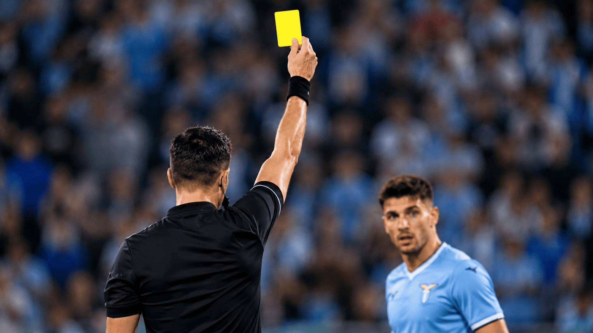 Referee holding up a yellow card during a professional soccer match while a player reacts on the field