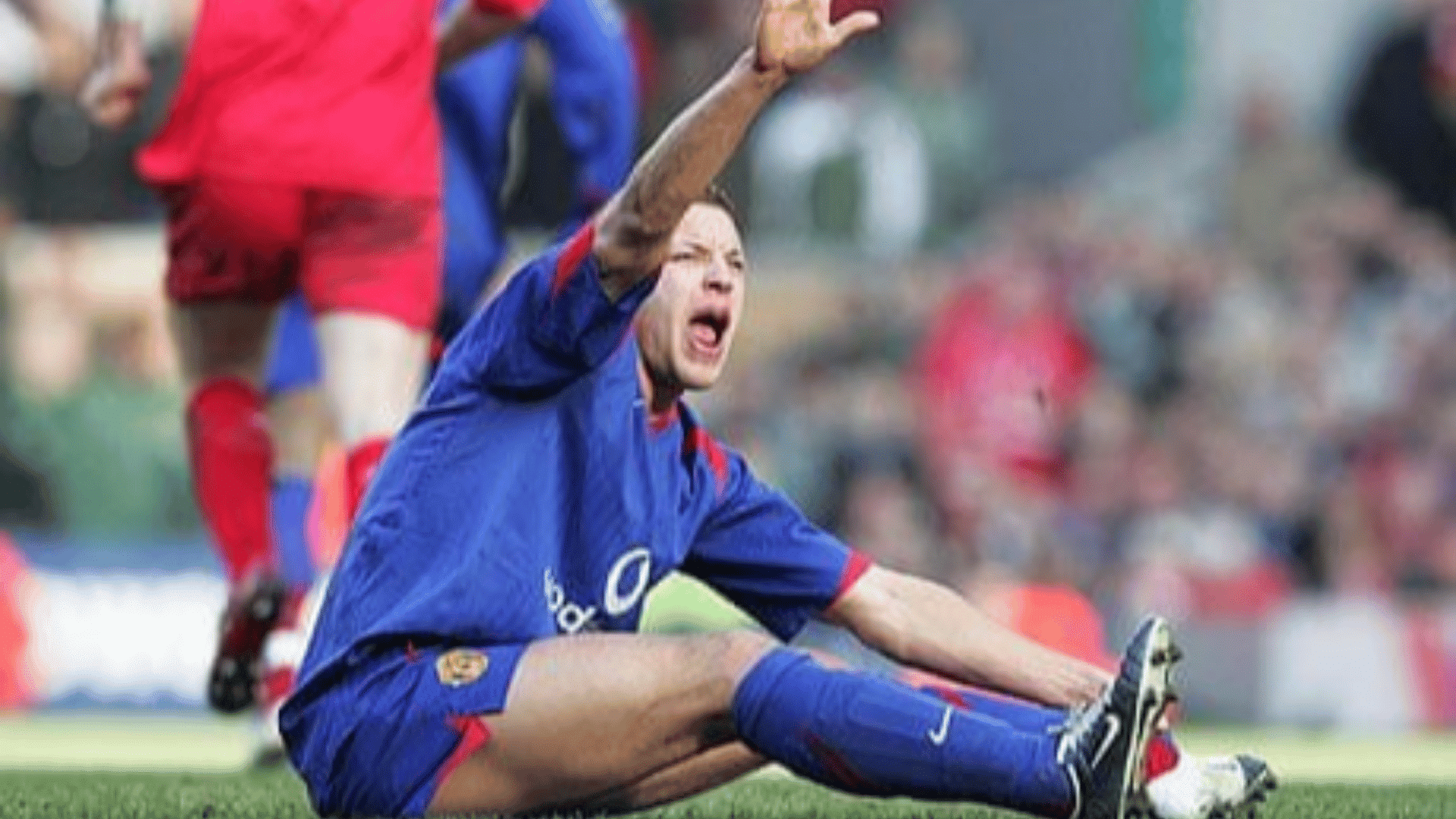 a football player in a blue kit sits on the grass raising one hand after a challenge during a match in a stadium