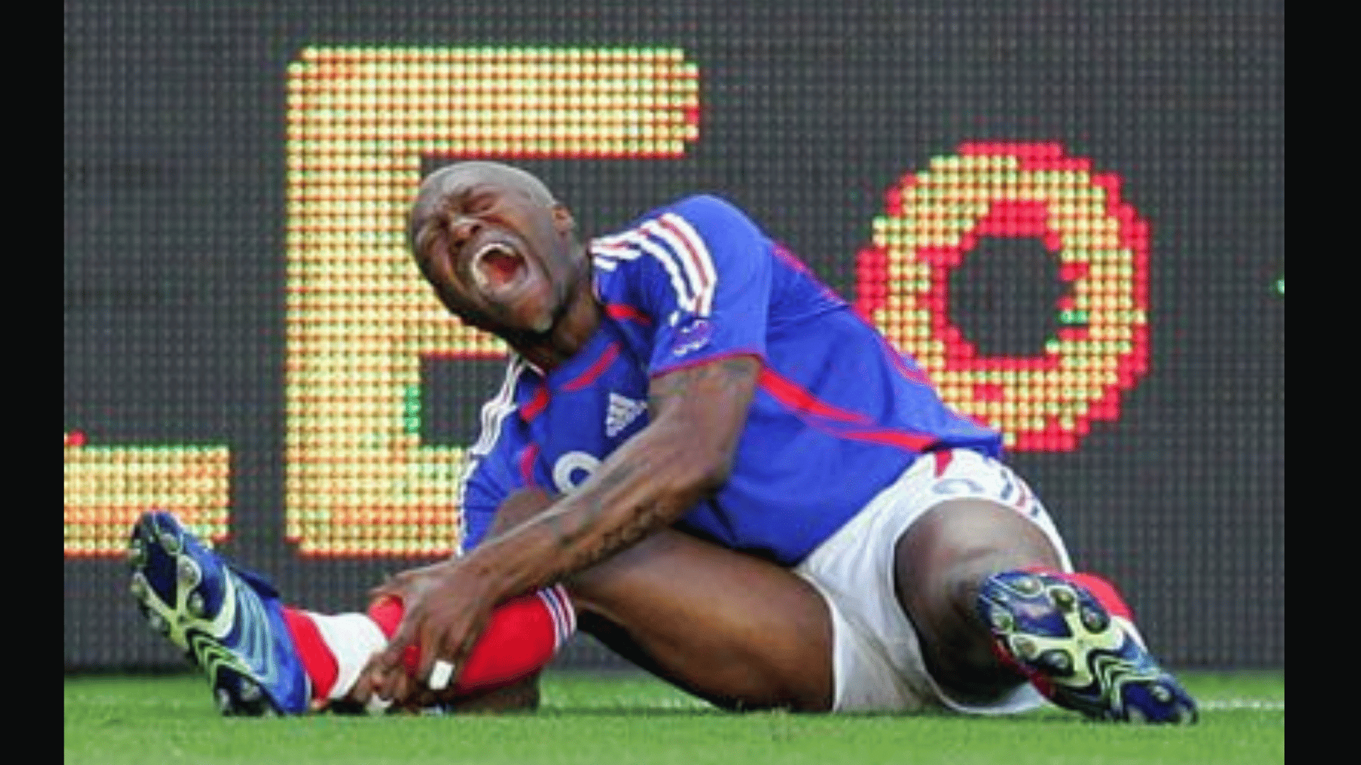 a football player in a blue kit sits on the pitch holding his leg and shouting in pain near a bright advertising board