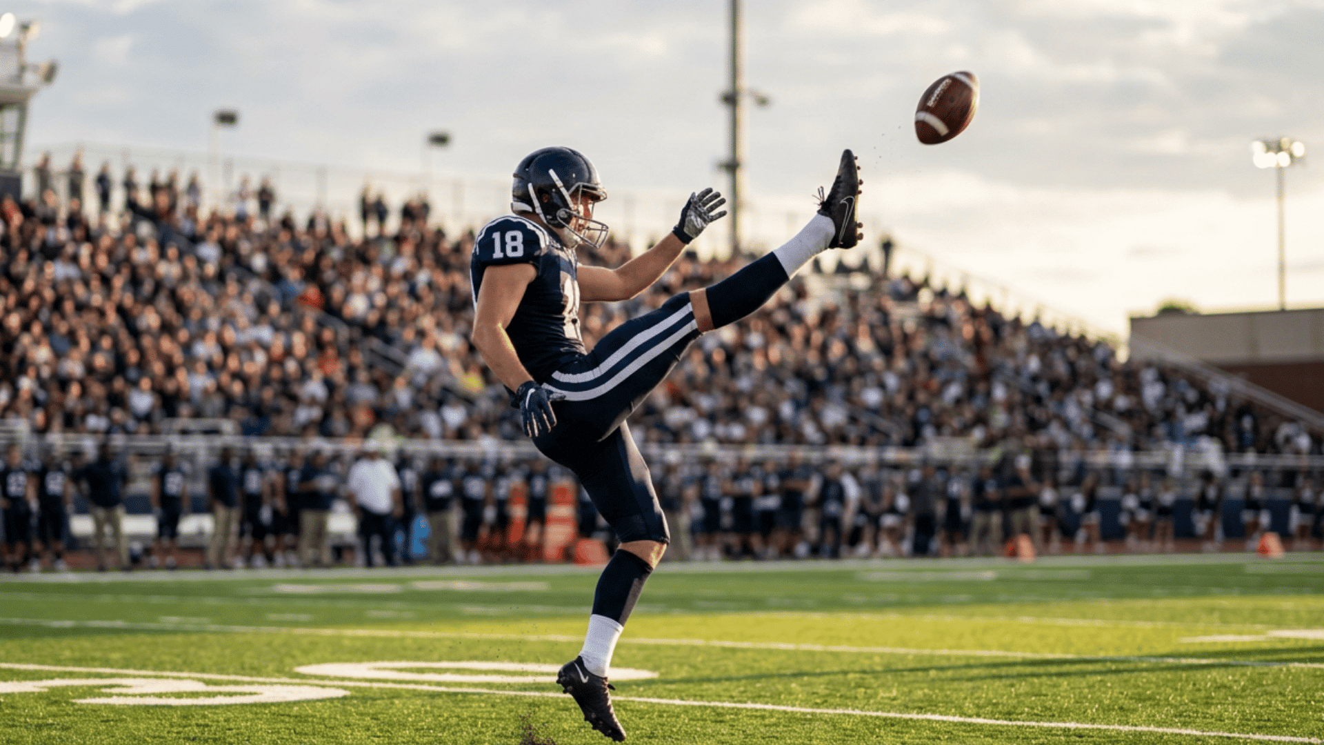 a football player in a navy uniform punts the ball on a field with a crowded stadium behind him