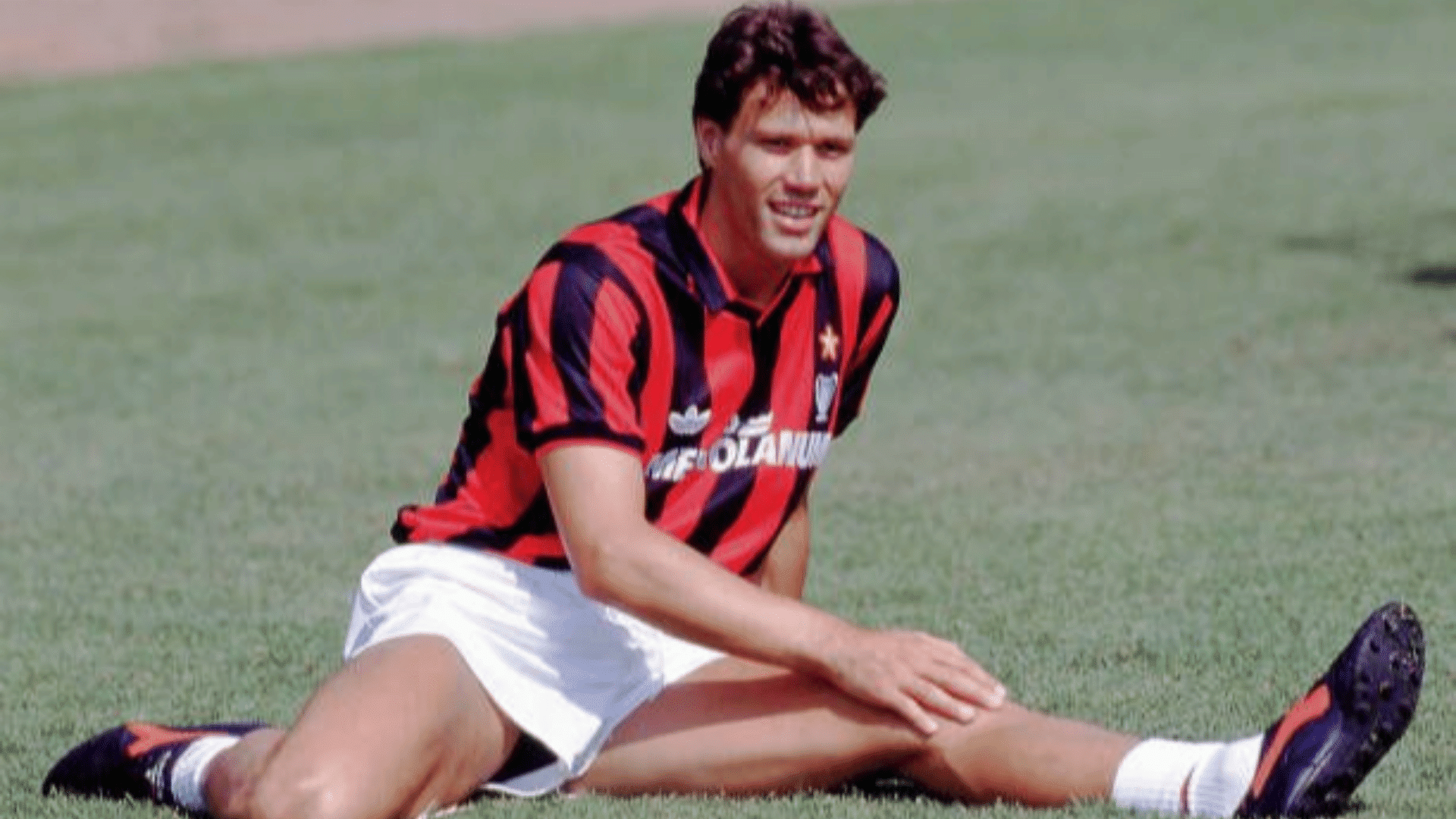 a football player in a red and black striped kit sits on the grass stretching one leg after injury
