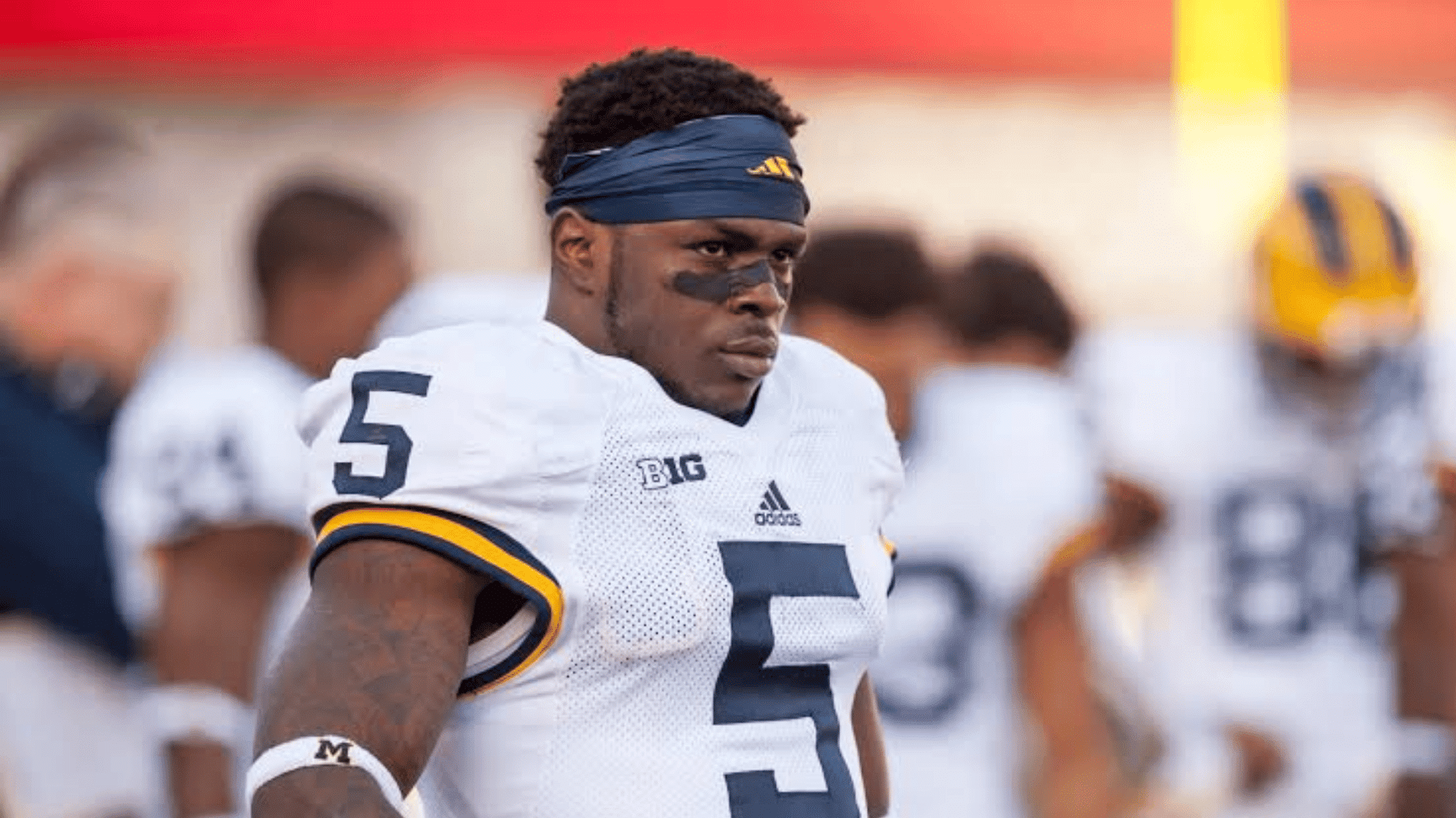 a football player in a white number 5 michigan jersey wearing a navy headband and eye black on the sideline
