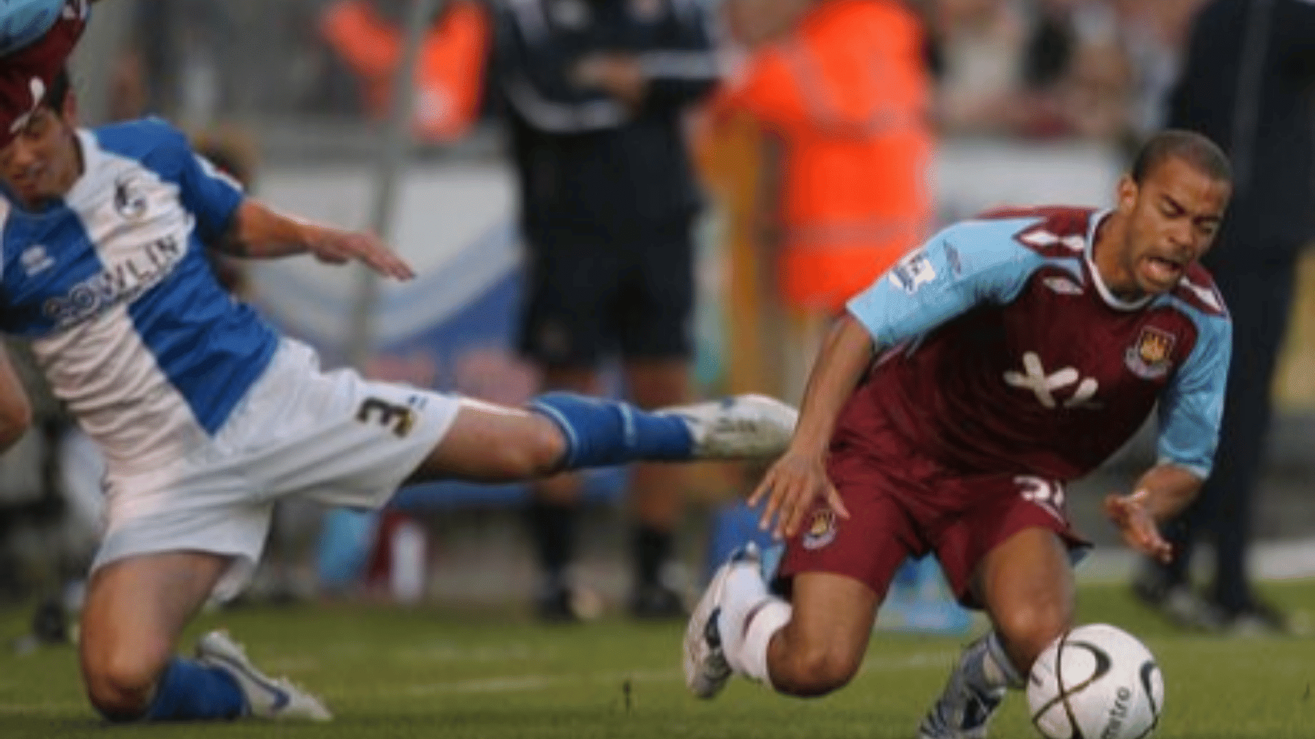 a football player in claret runs with the ball as an opponent in blue and white slides in with an outstretched leg
