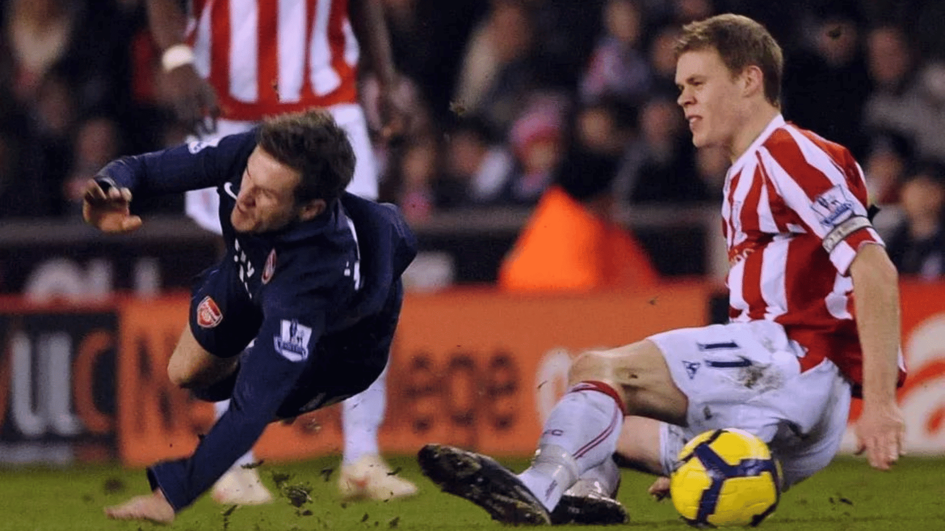 a football player in navy falls forward after a sliding tackle from an opponent in red and white stripes