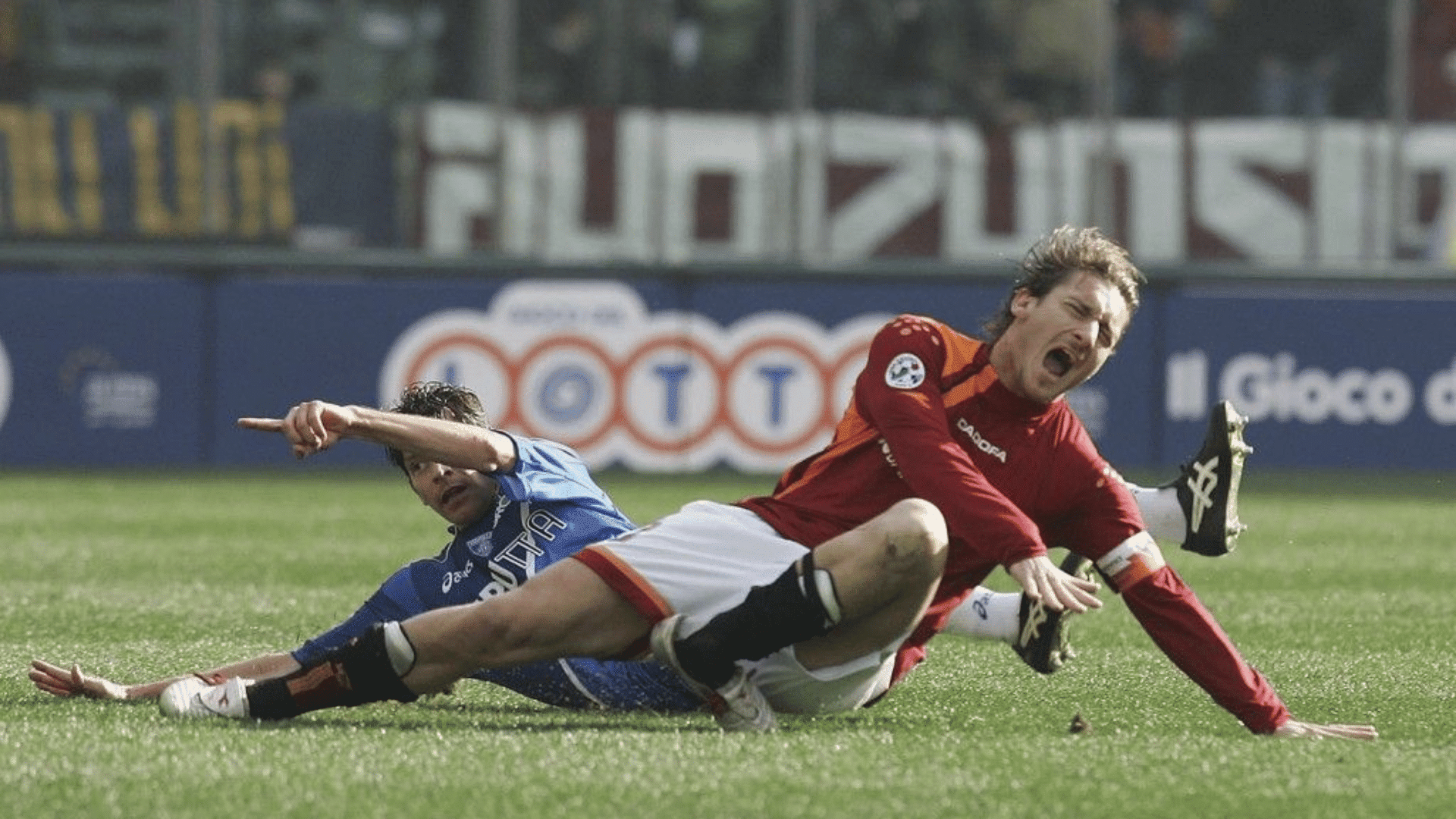 a football player in red is knocked down by a sliding opponent in blue during a tackle on a sunny pitch