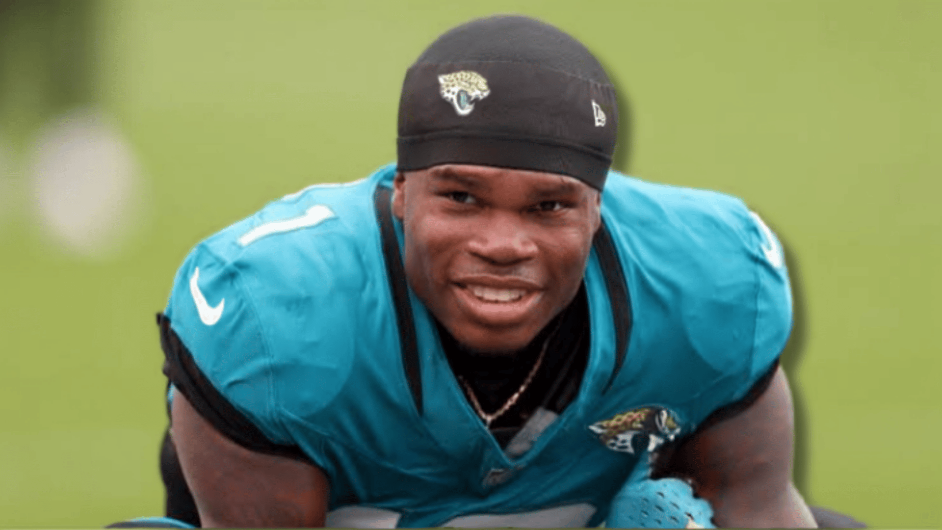 a jaguars football player in a teal jersey and black cap smiles while leaning forward on a green practice field