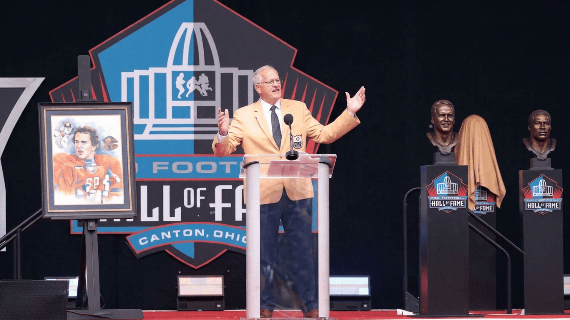 a man in a gold jacket speaks at a football hall of fame ceremony with portraits and busts on stage