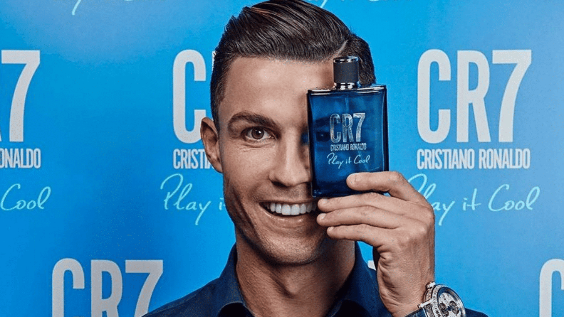 a smiling man holds a blue cr7 cologne bottle over one eye in front of a bright blue fragrance backdrop