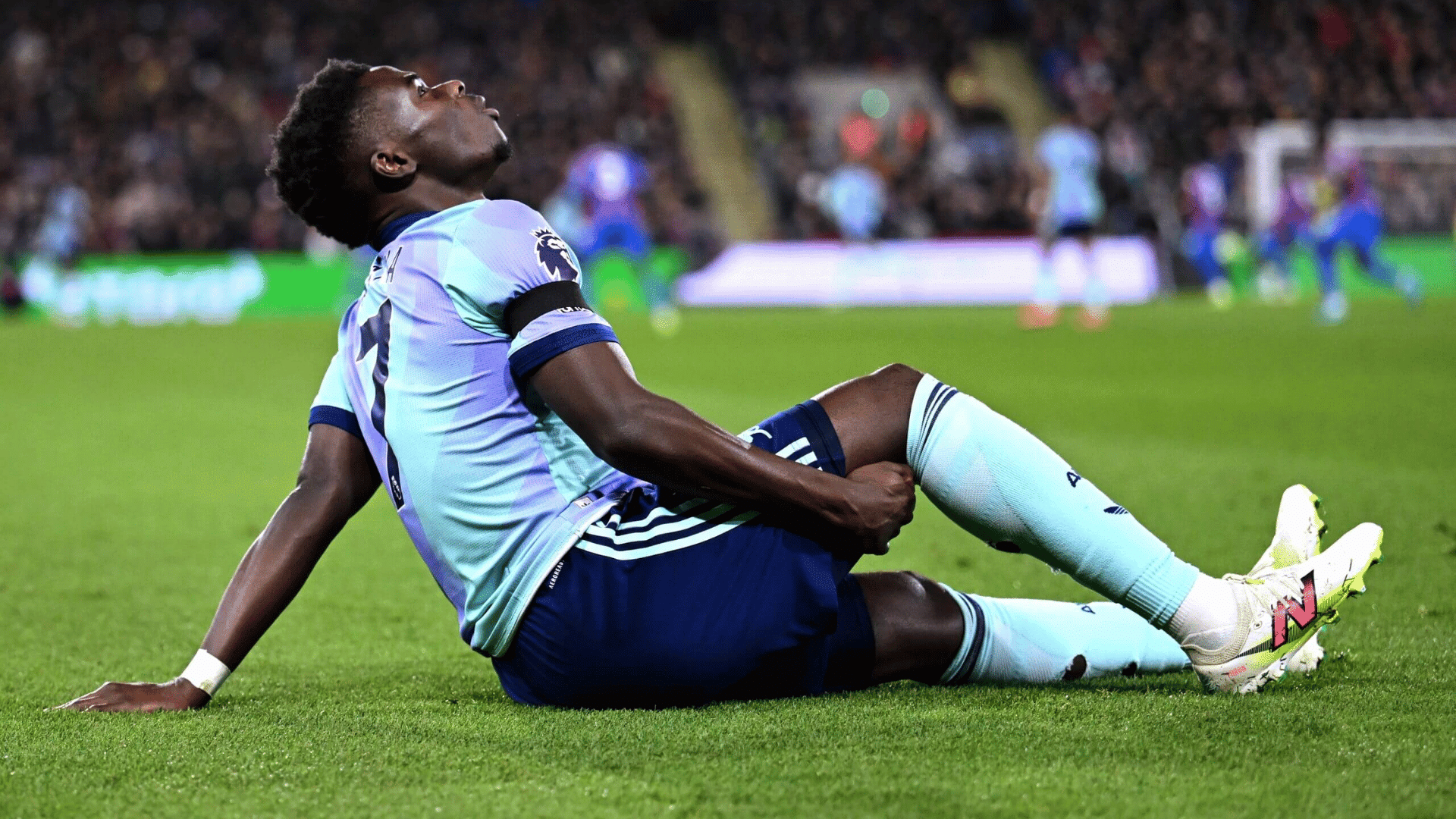a soccer player in a light blue kit sits on the grass clutching his leg with blurred players and crowd behind him