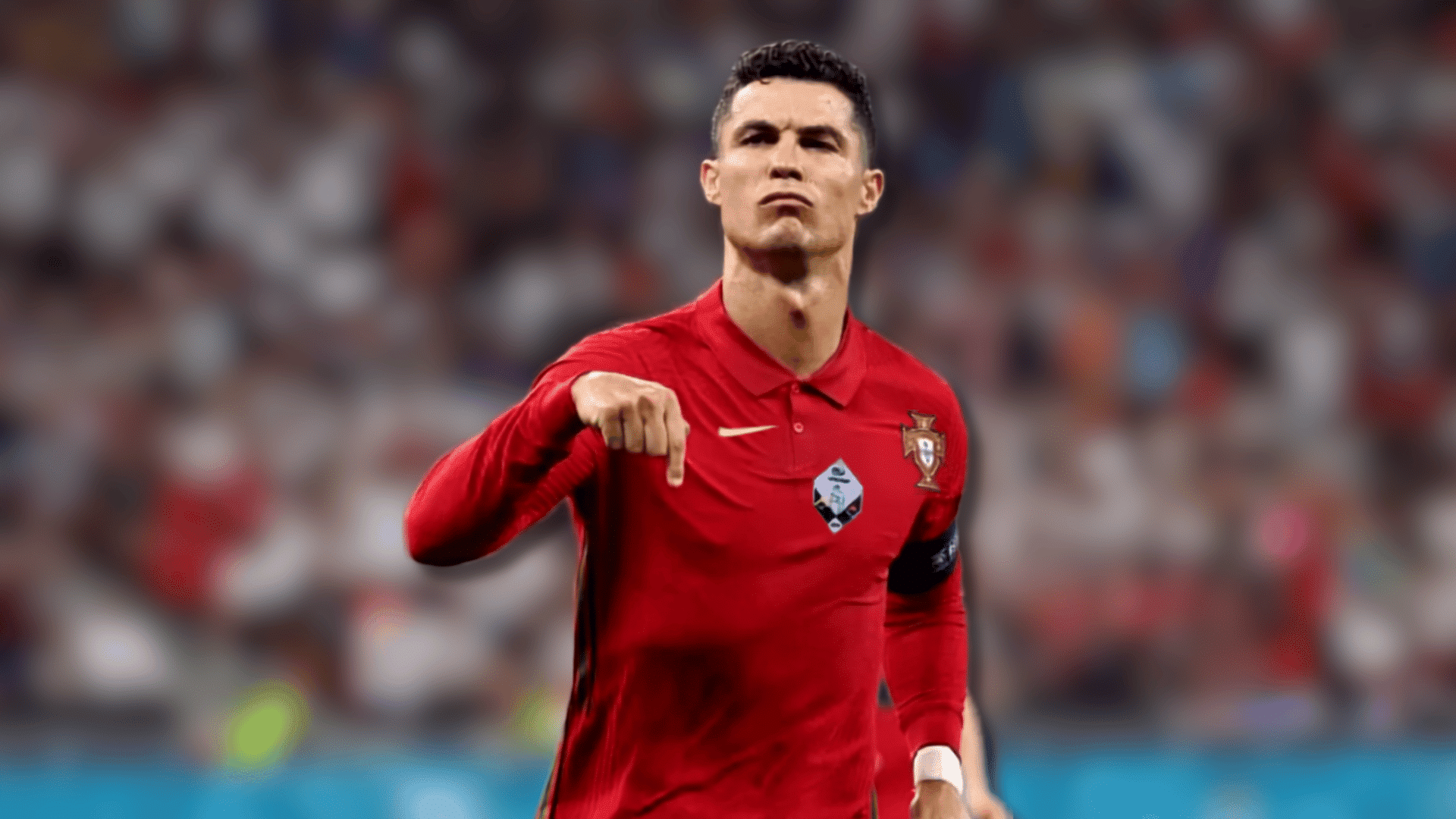 a soccer player in a red jersey points downward with a stern expression as a blurred crowd fills the stadium