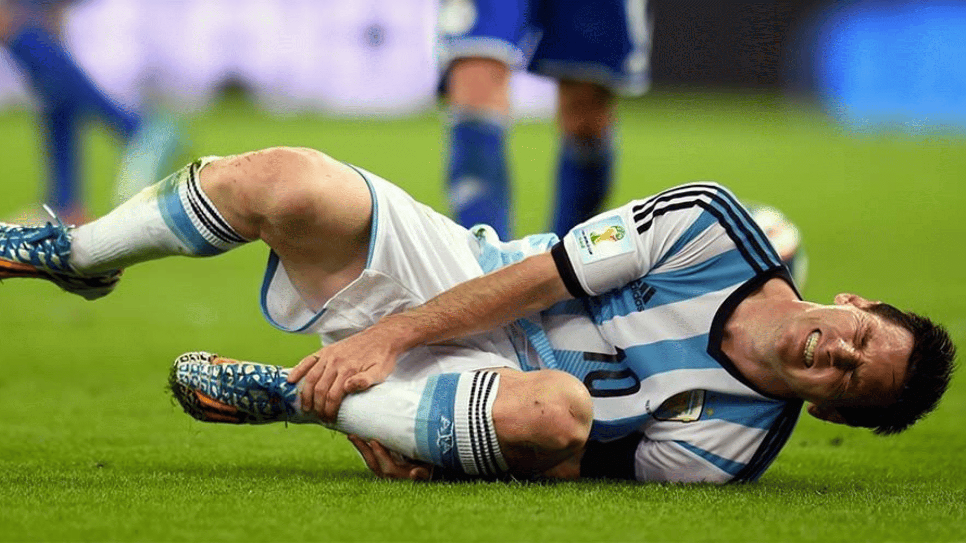 a soccer player in blue white kit lying on grass holding his ankle in pain during a game while another player approching