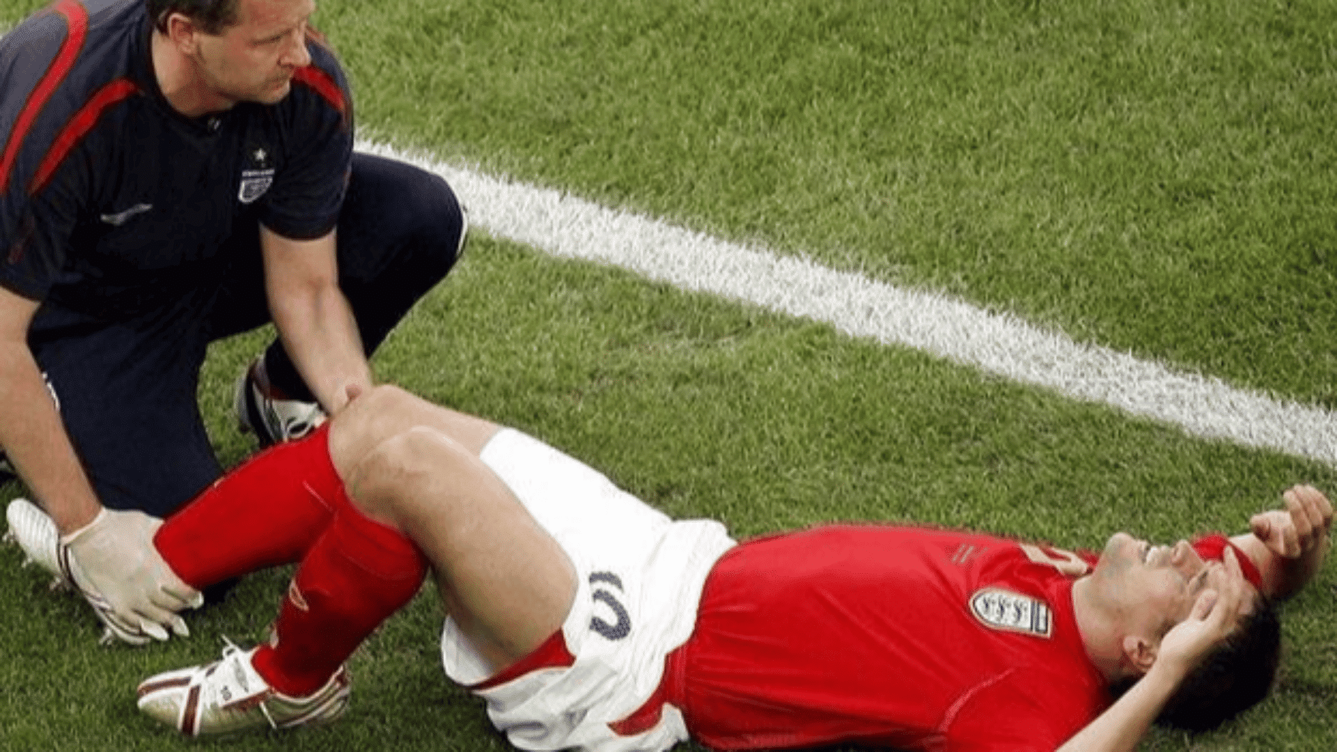 a trainer checks a football players leg as he lies on the grass holding his head after an injury during a match