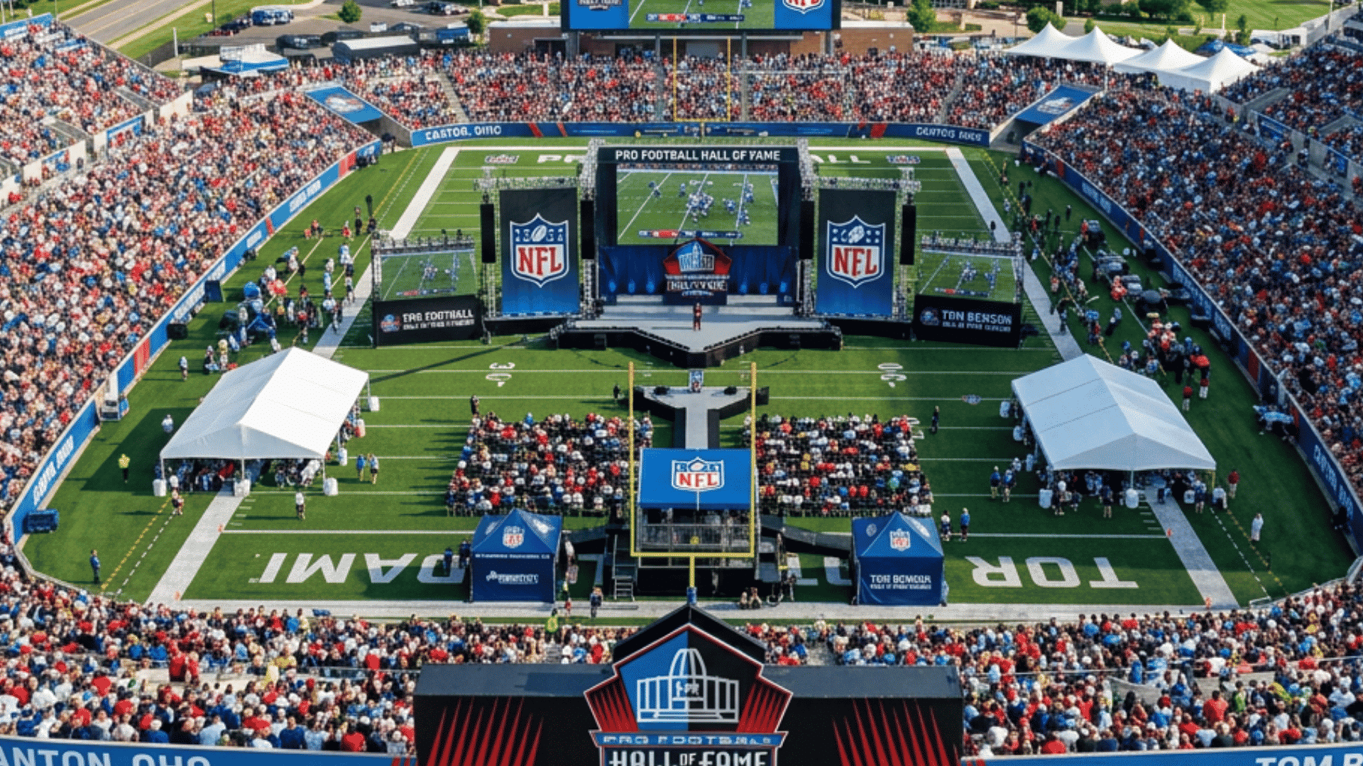 aerial view of a crowded football stadium with hall of fame banners stage screens roads and trees around it