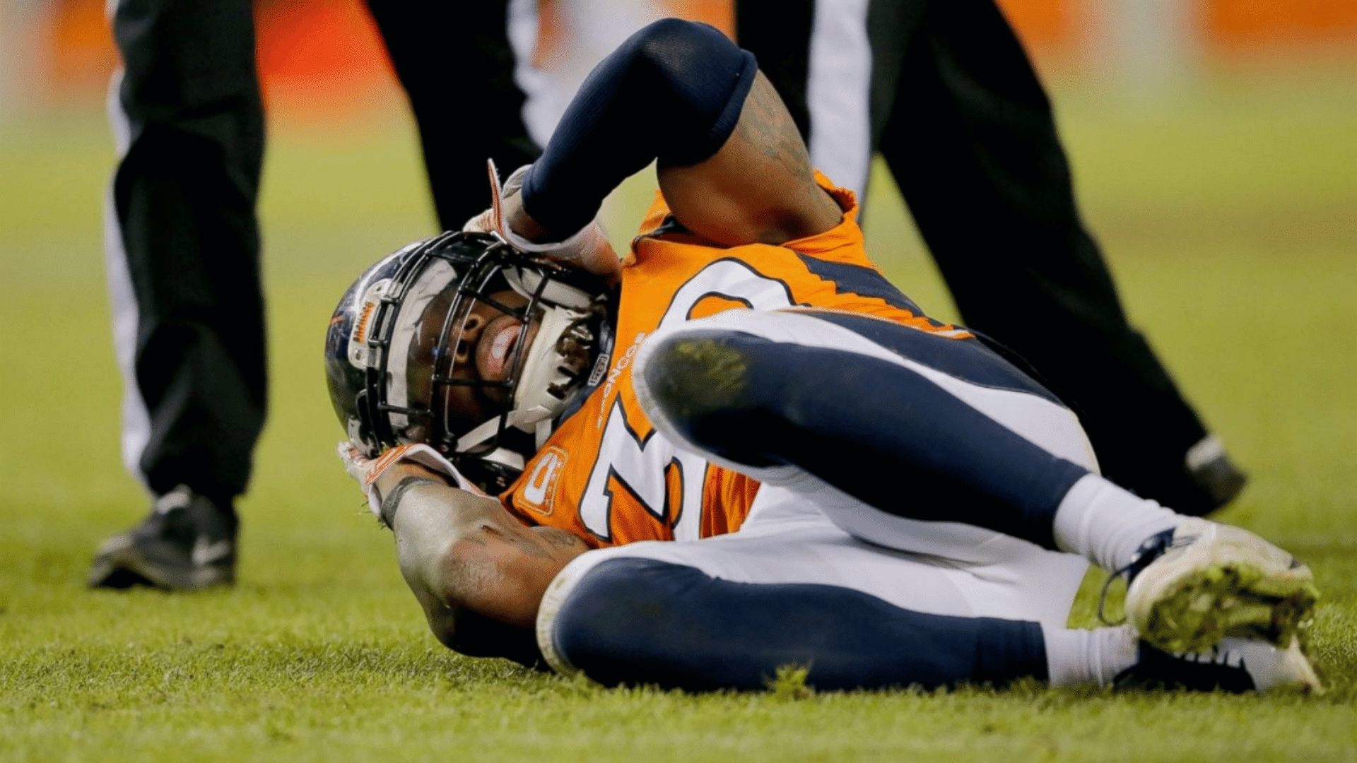 american football player in orange jersey lying on grass clutching head in pain during stadium match with helmet on