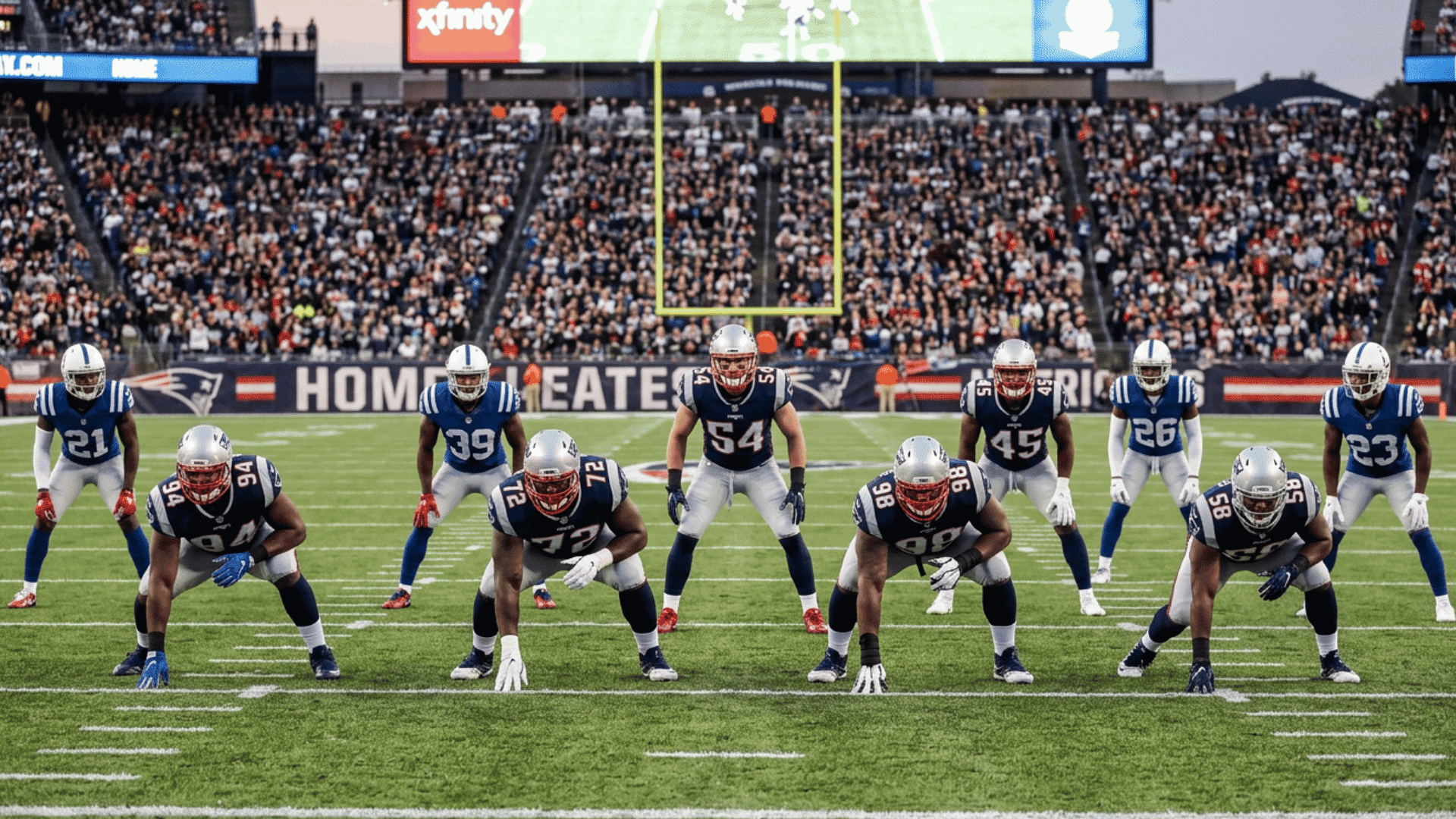 american football team in blue and white uniforms lined up in defensive stance on field in stadium with crowd and scoreboard