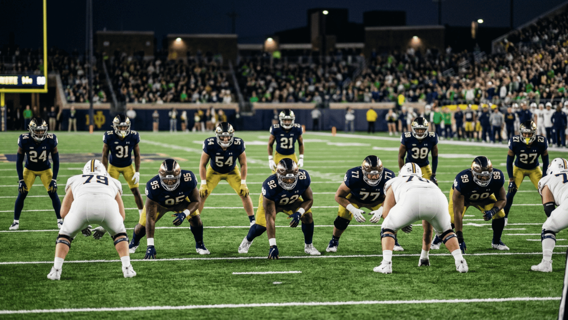 american football teams lined up at line of scrimmage during night game in stadium with crowd in background