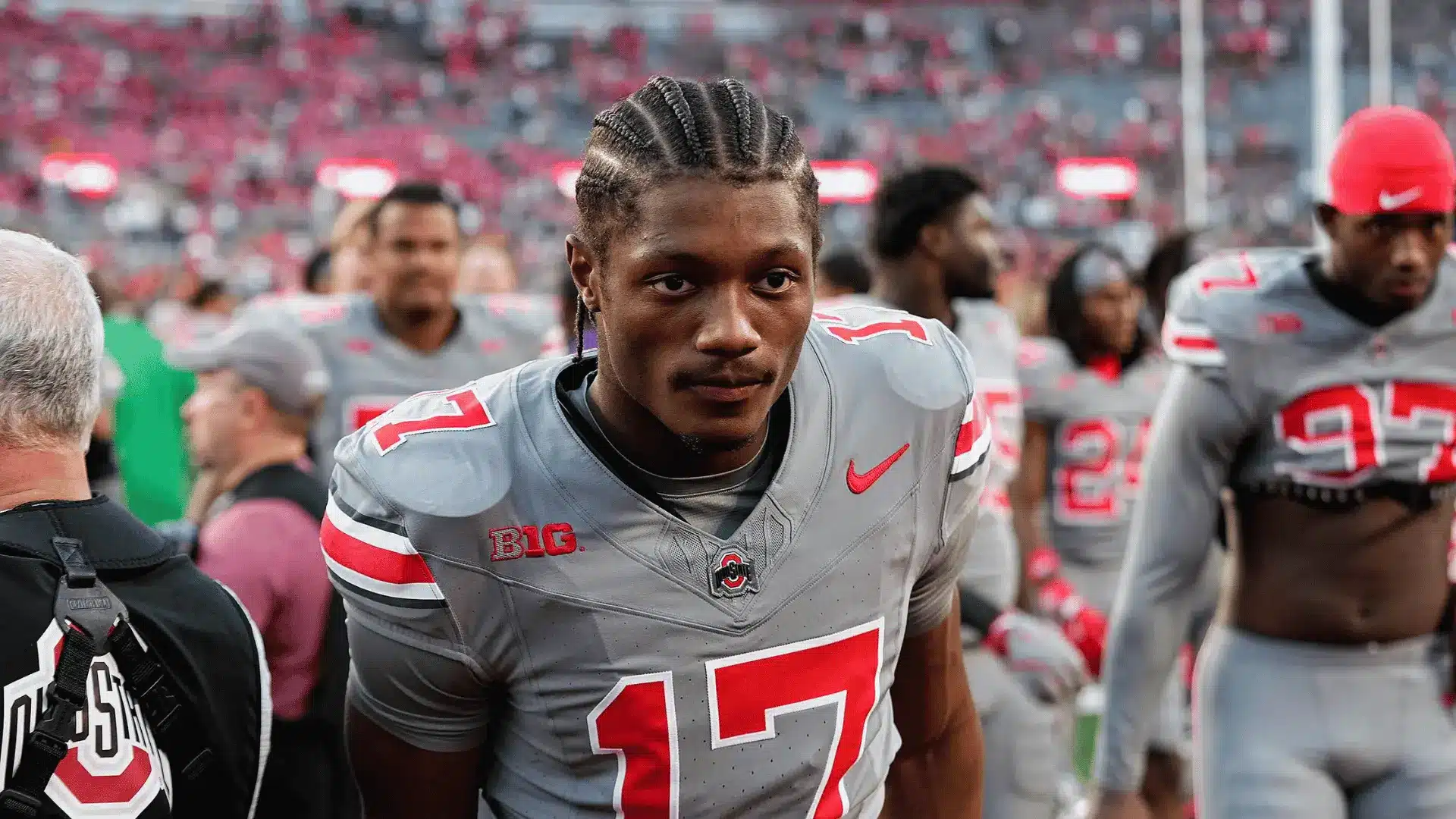 football player in a gray ohio state jersey stands on the sideline with teammates behind him in a stadium