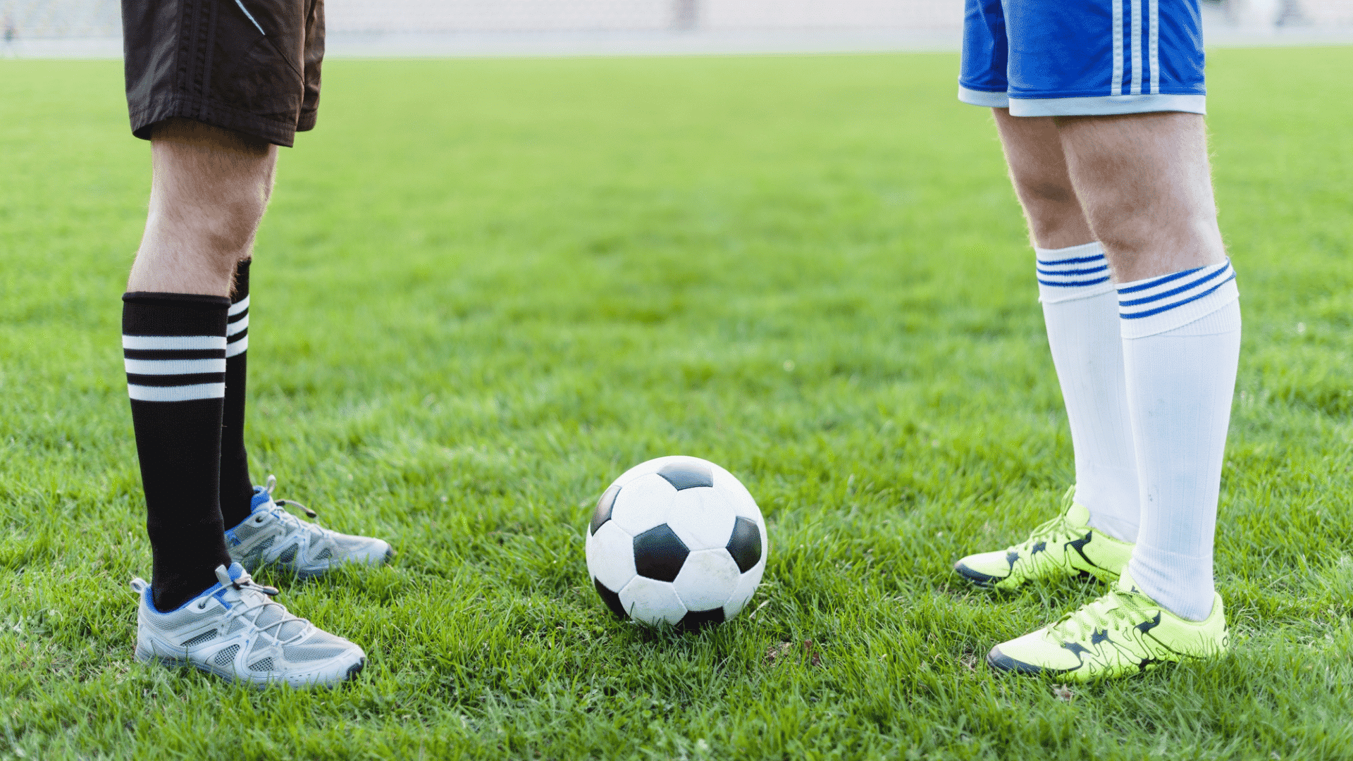 close up of two football players legs standing on green grass field facing each other with a standard black and white ball
