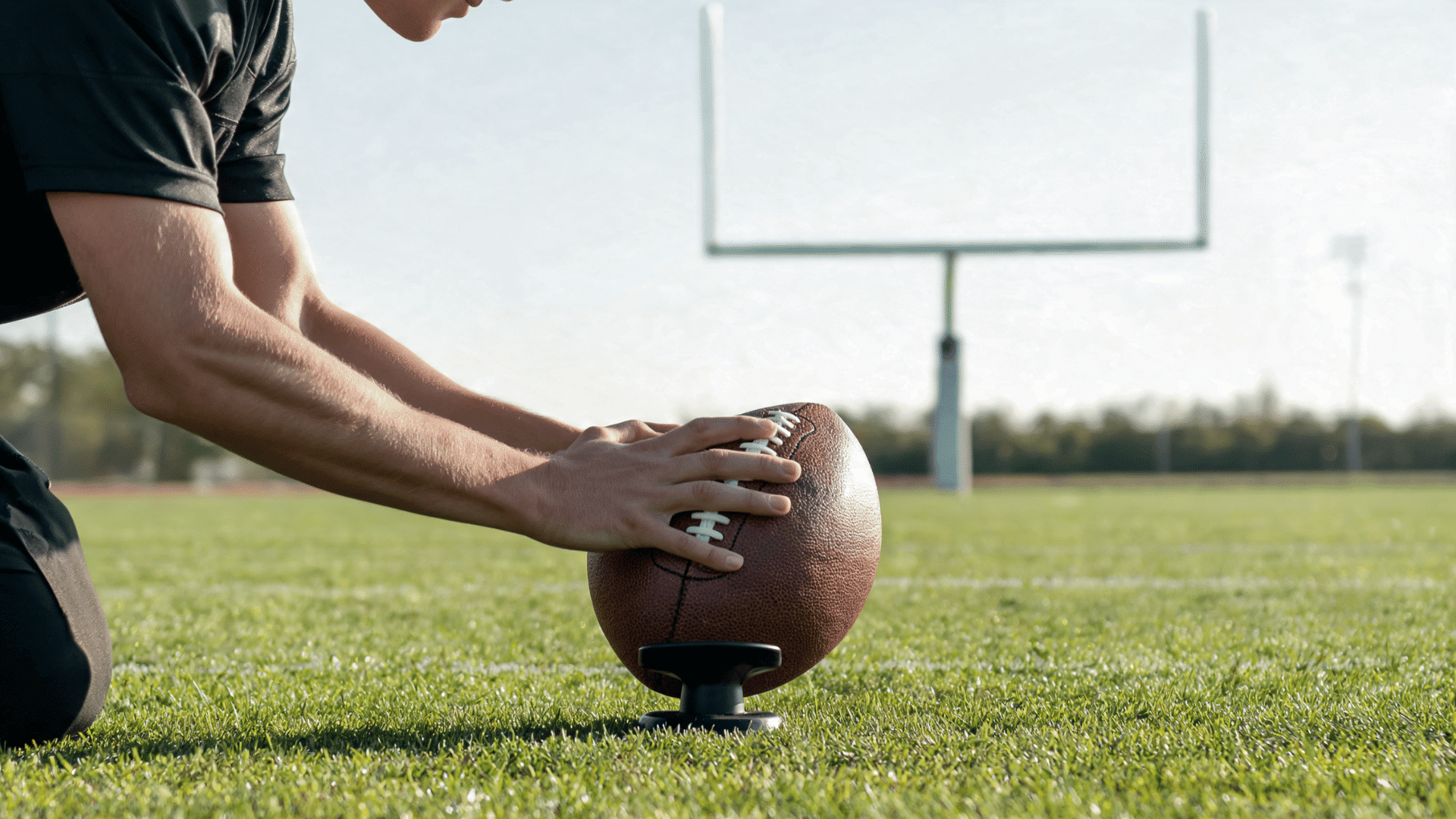 close view of a beginner placing an american football securely on a low tee while aiming toward the goalposts