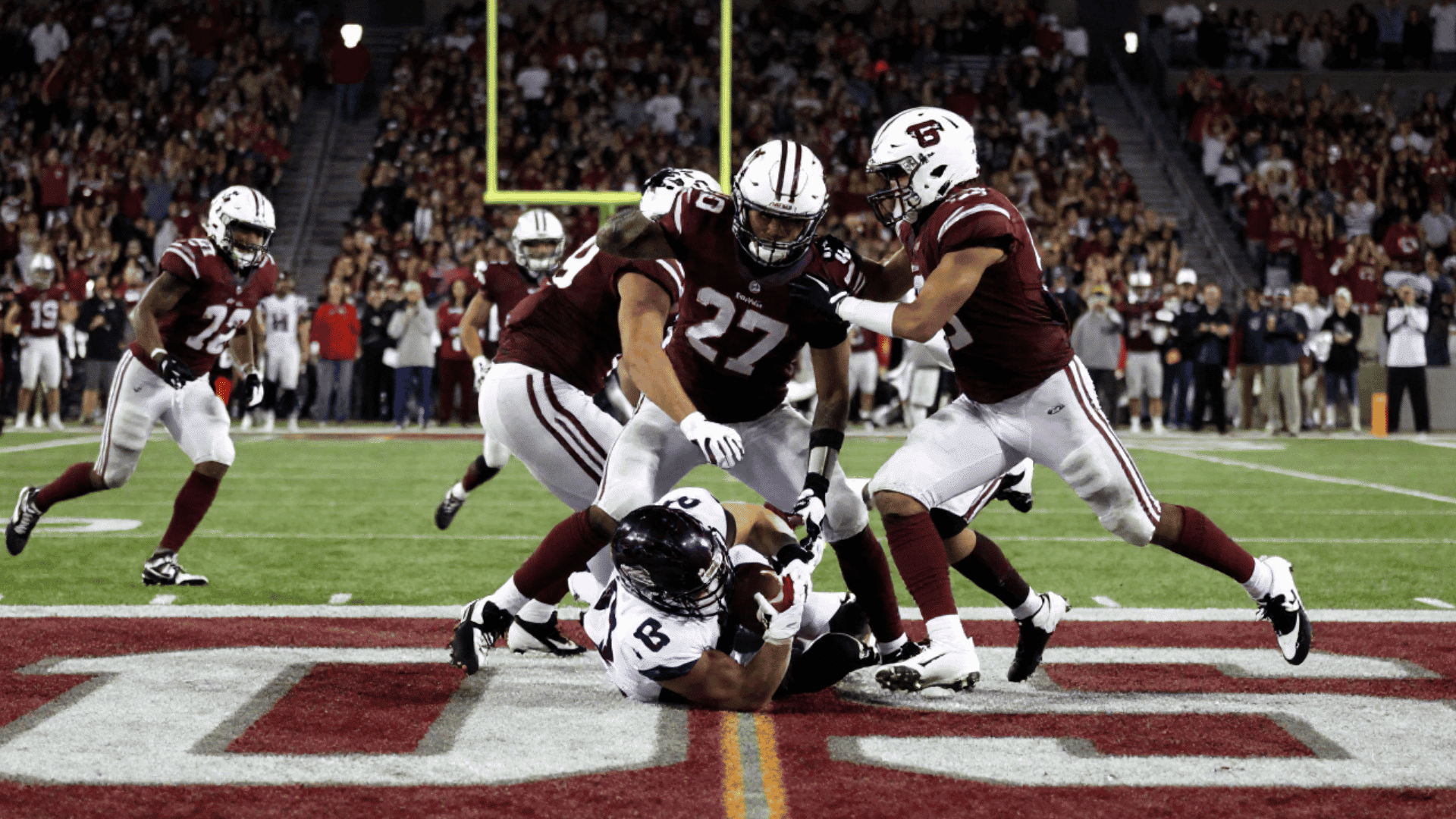 football player in a white and purple uniform is tackled on the ground by multiple players in maroon and white uniforms