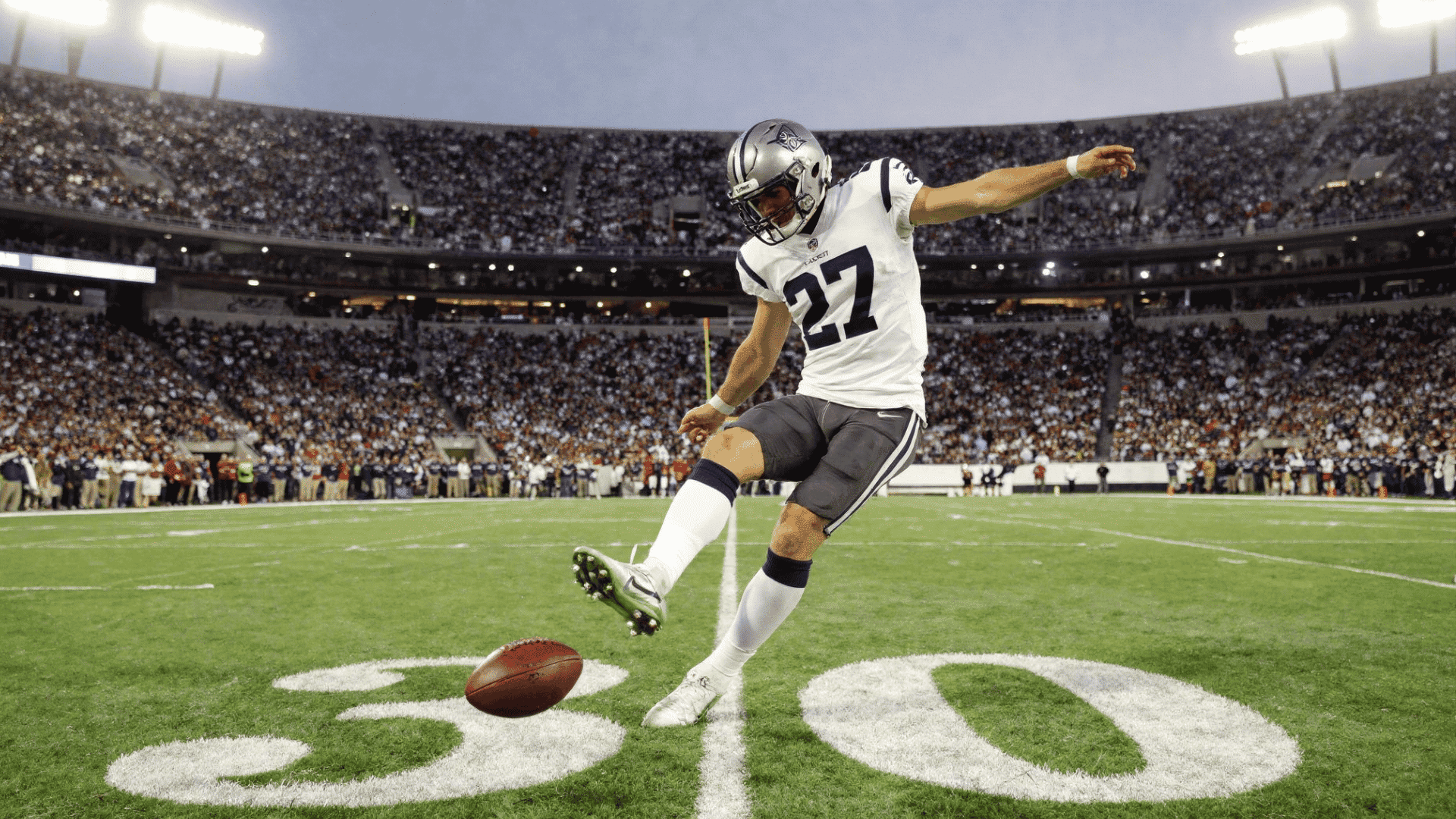 football player in a white uniform kicks a football on a field in front of a stadium full of spectators
