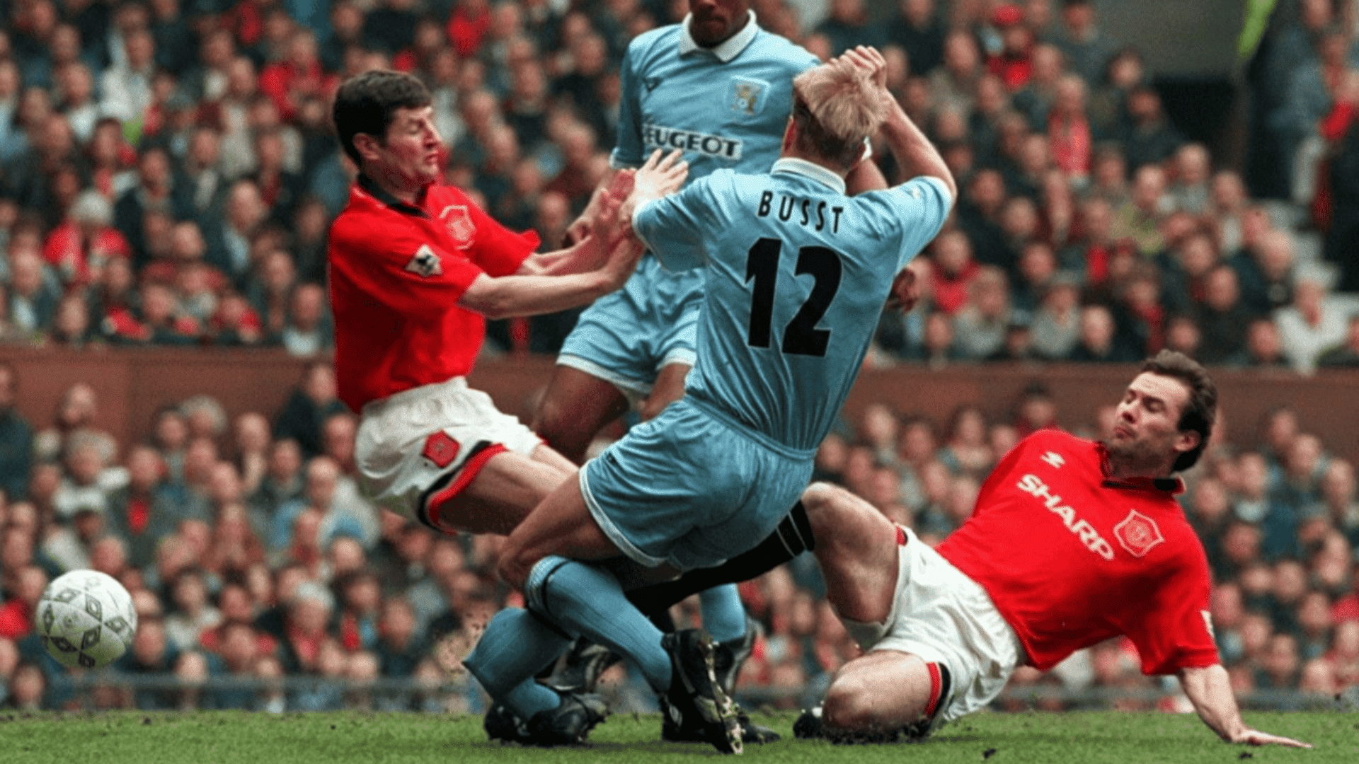 football players in red and blue kits collide during a tackle as the ball rolls across a crowded stadium pitch