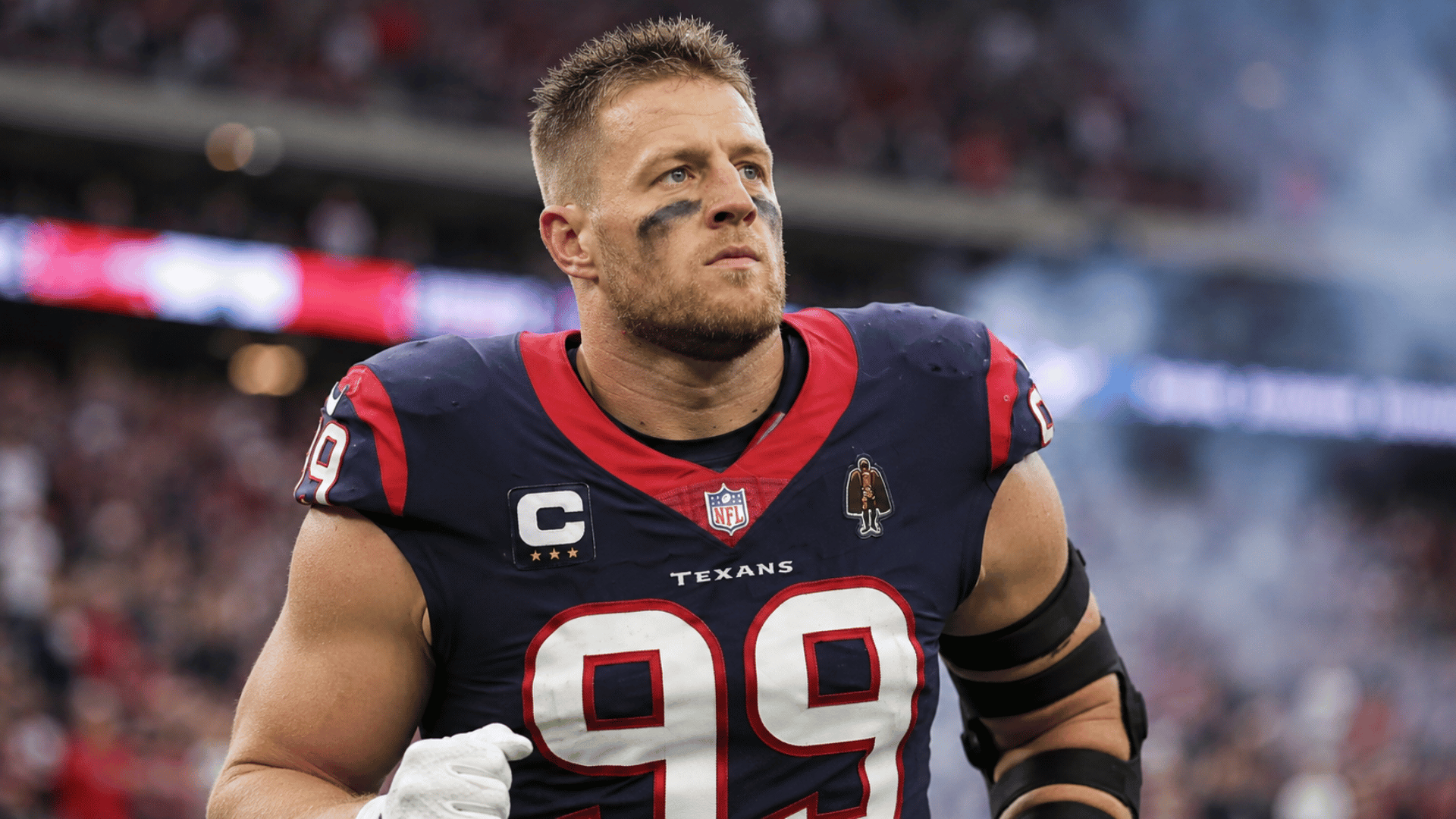 j j watt standing on the field after a game with stadium lights and a blurred crowd behind him