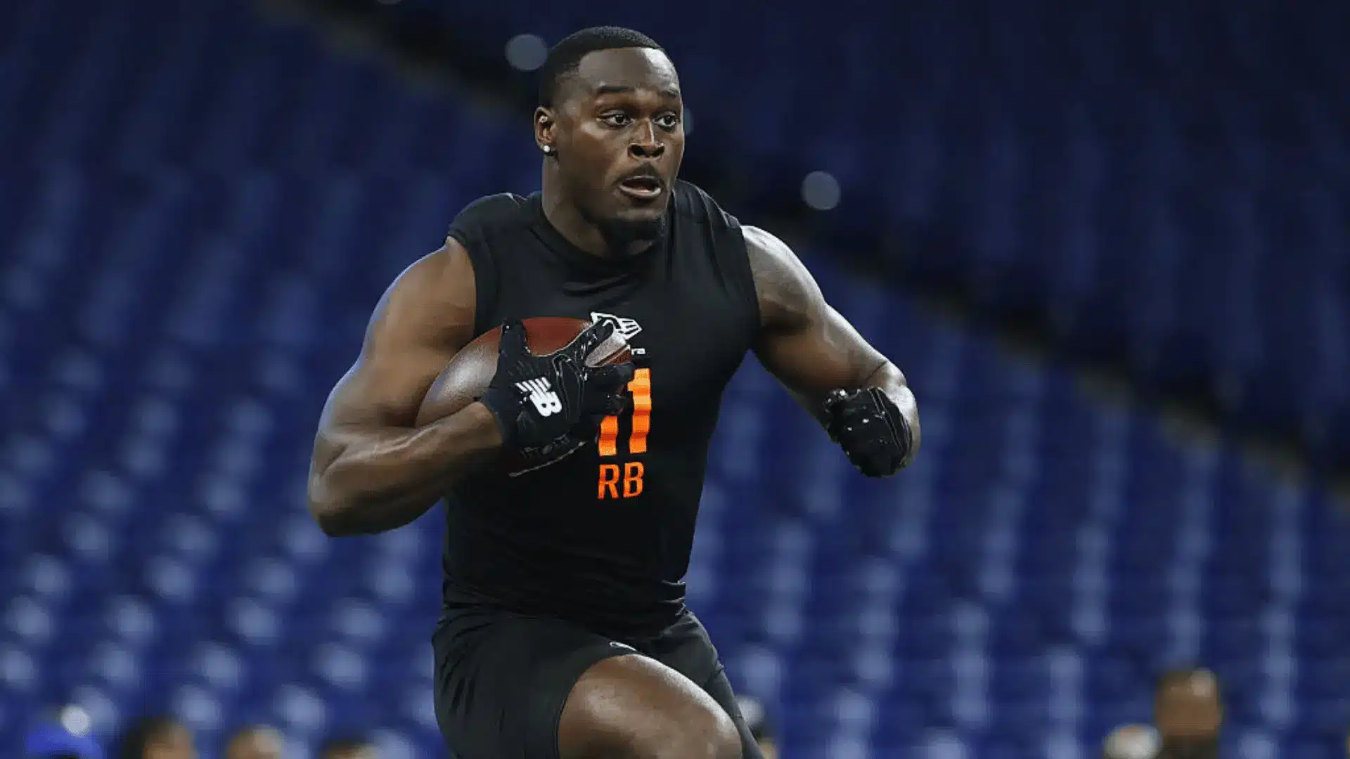 football player in black workout gear runs with a ball during drills inside a stadium with blue seats