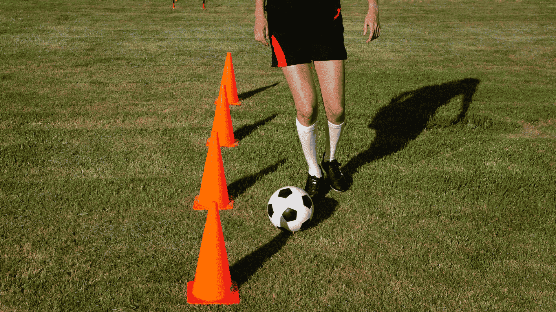 legs of an athlete wearing black cleats dribbling a football closely around bright orange training cones on green grass