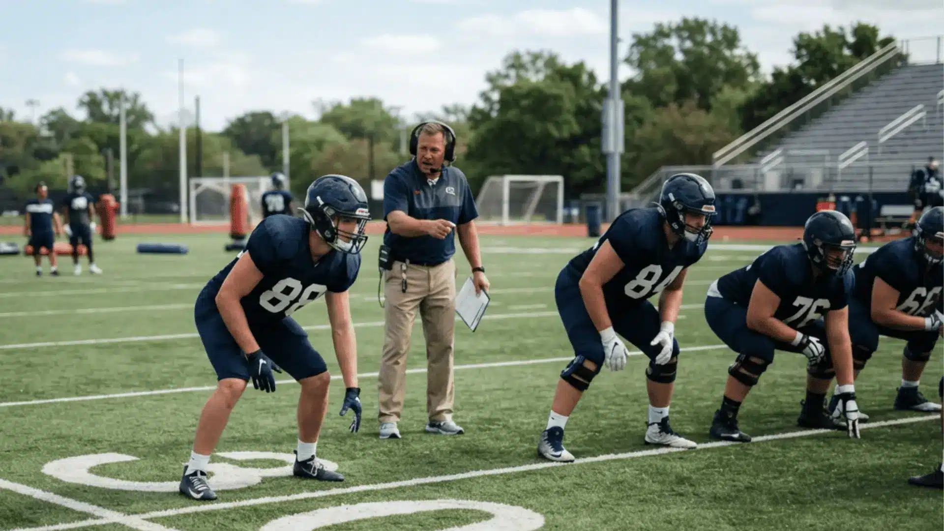 football players line up for a drill while a coach with a headset points across a green practice field