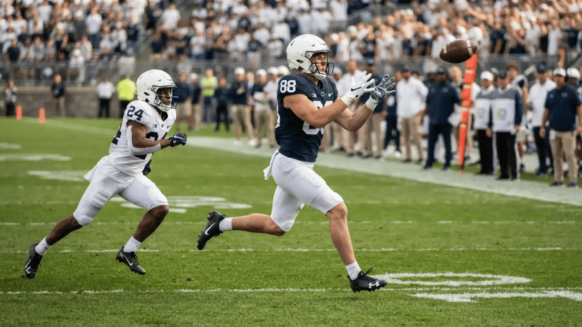 a football receiver in a dark jersey runs with his hands out for a pass as a defender in white chases behind