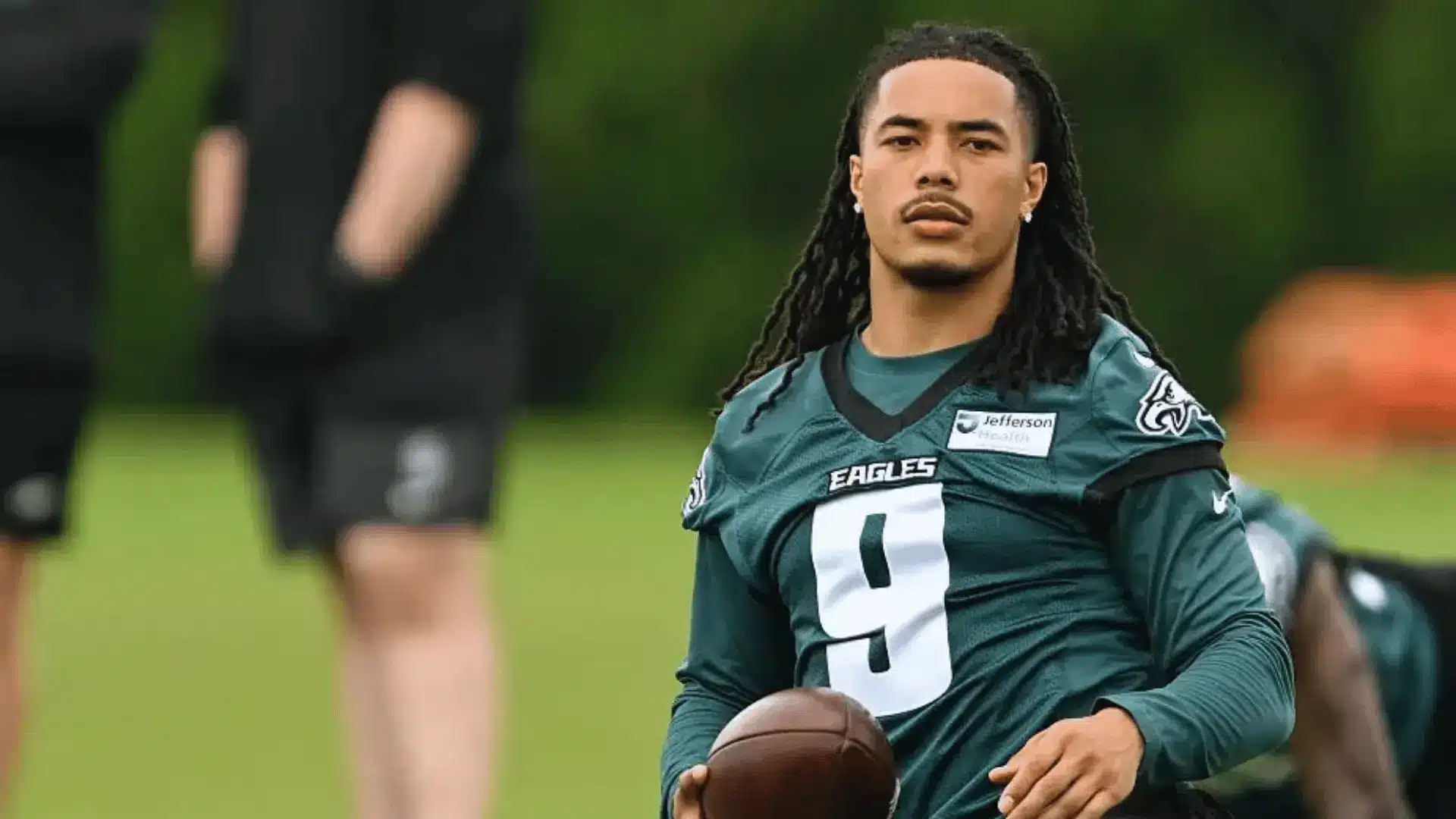 football player in a green eagles practice jersey holds a ball while walking on a field during team drills