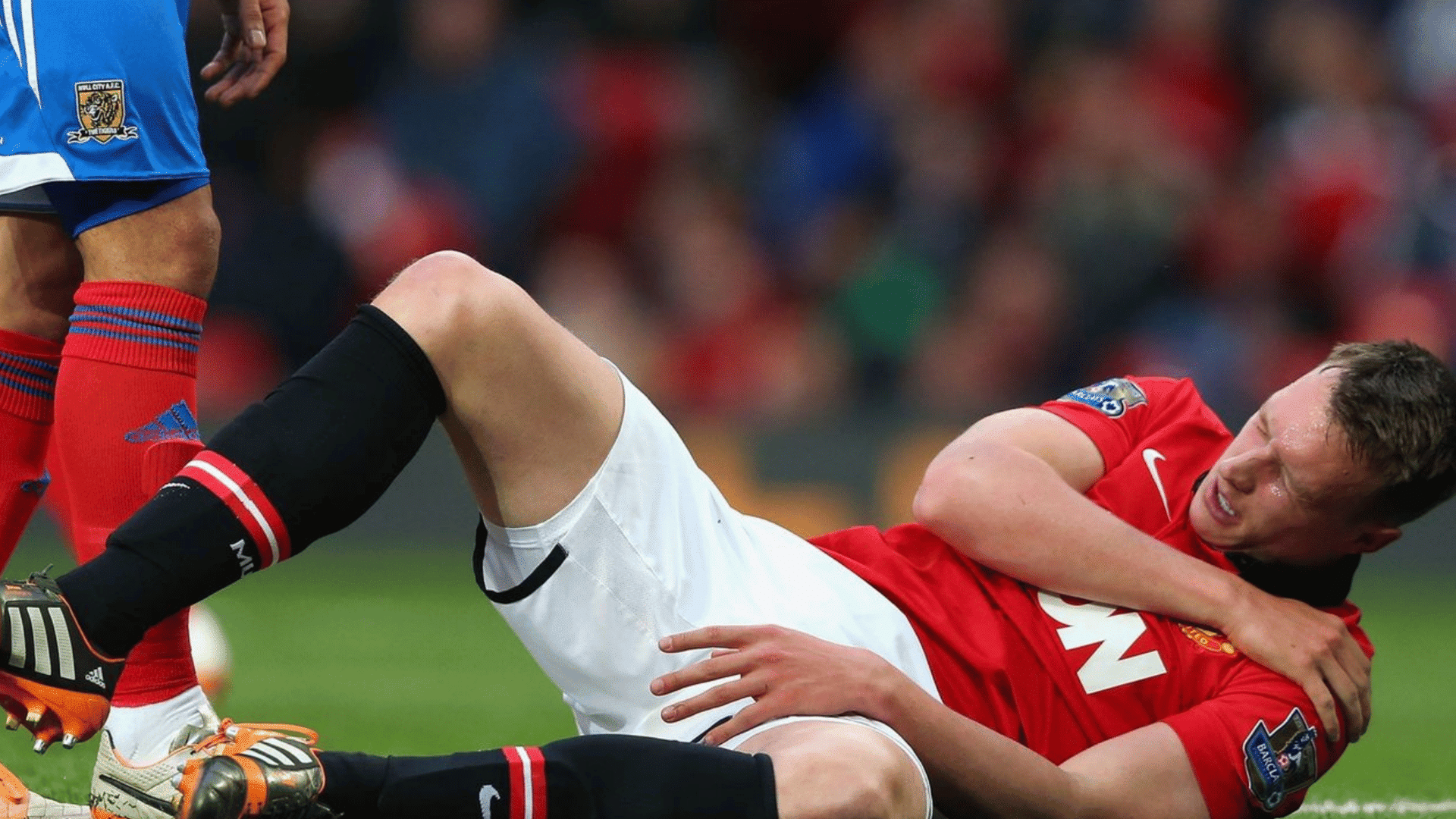 male soccer player in red jersey lying on grass clutching his shoulder in pain grimacing during match stadium background