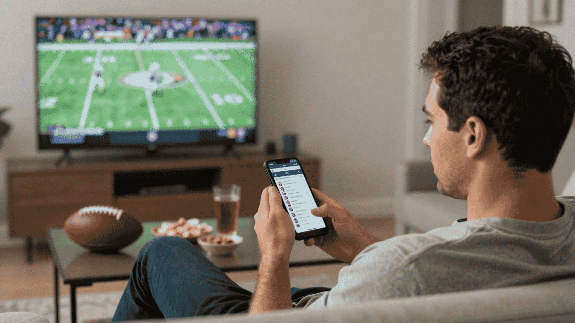 man checks fantasy football lineup on a phone while watching a football game on television at home