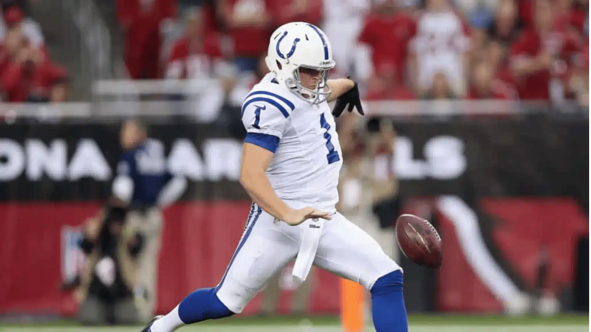 an american football player in a white and blue uniform punts the ball on the field before a blurred crowd in red