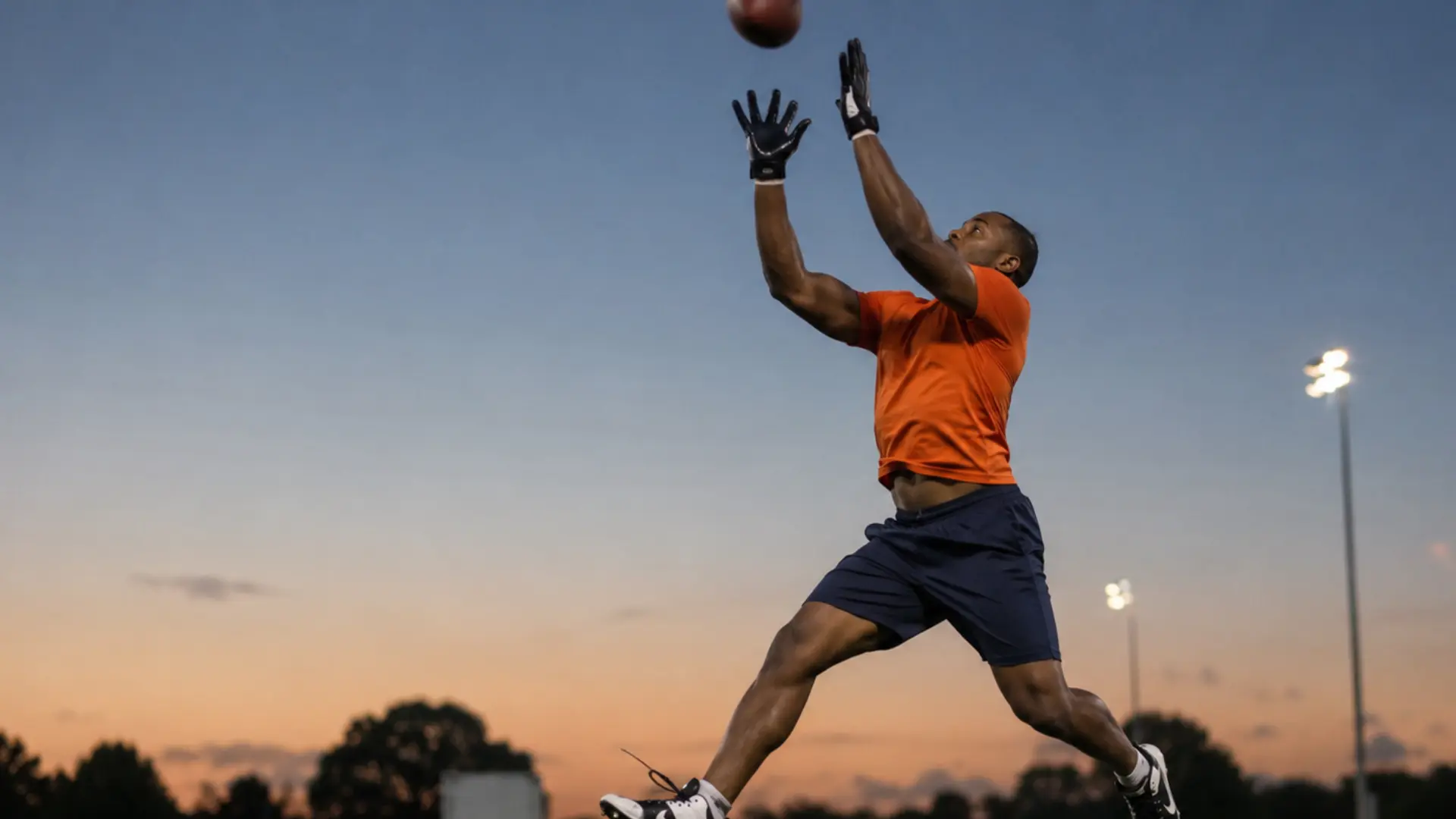 player stretches upward to catch a football against a glowing dusk sky during practice
