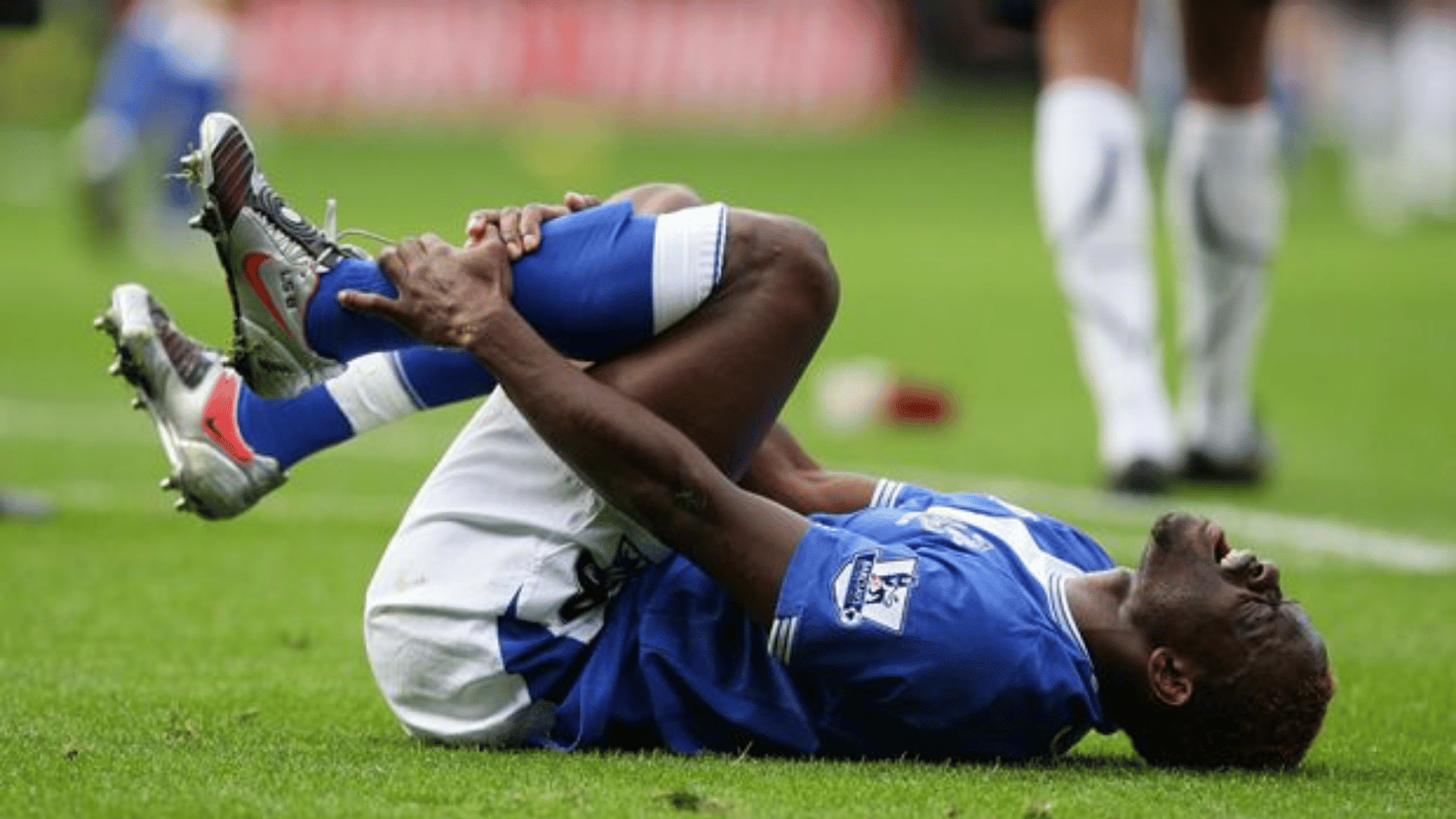 professional soccer player in blue kit lying on grass clutching knee in pain during stadium match