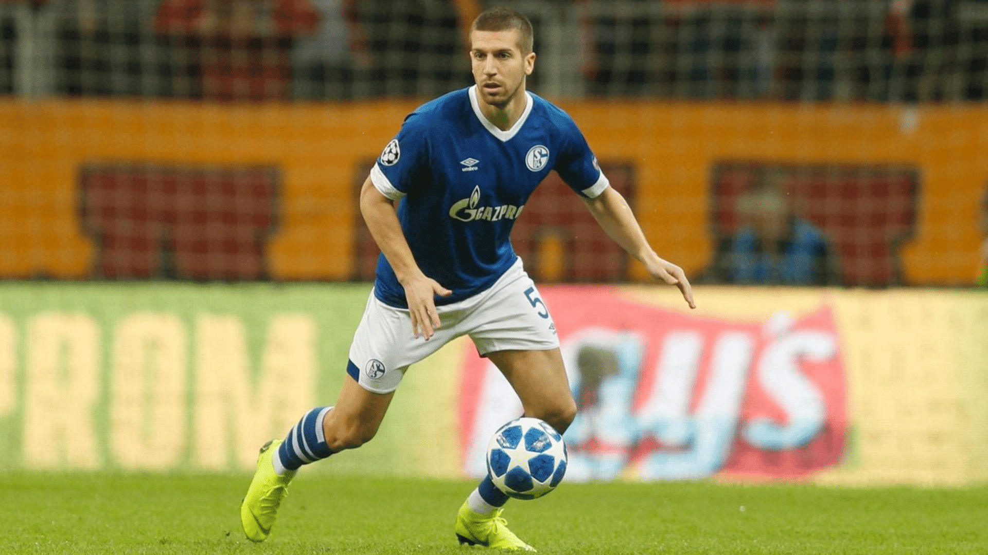 soccer player in a blue uniform dribbles the ball on the field during a match with stadium seats behind him