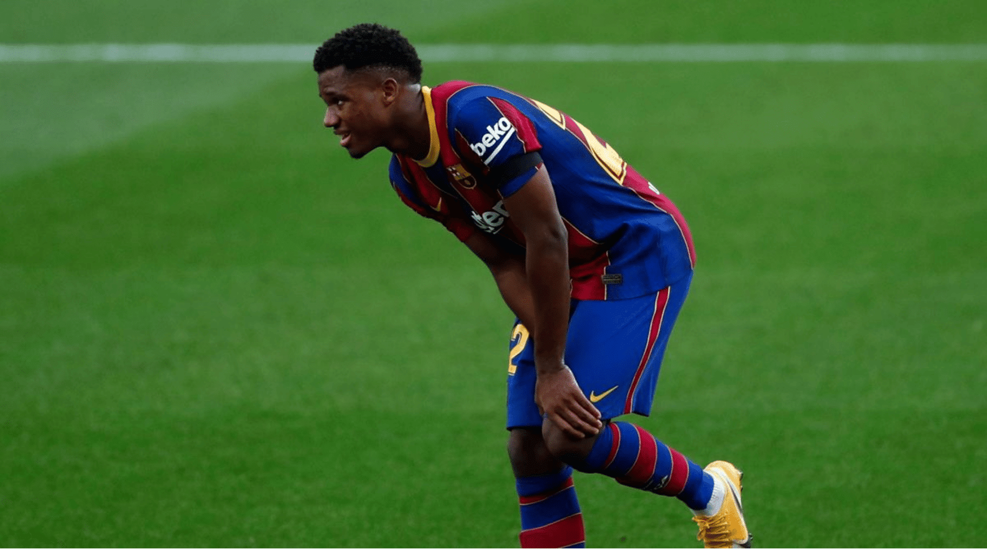soccer player in blue and red barcelona kit bending forward holding his knee on green pitch during match looking fatigued