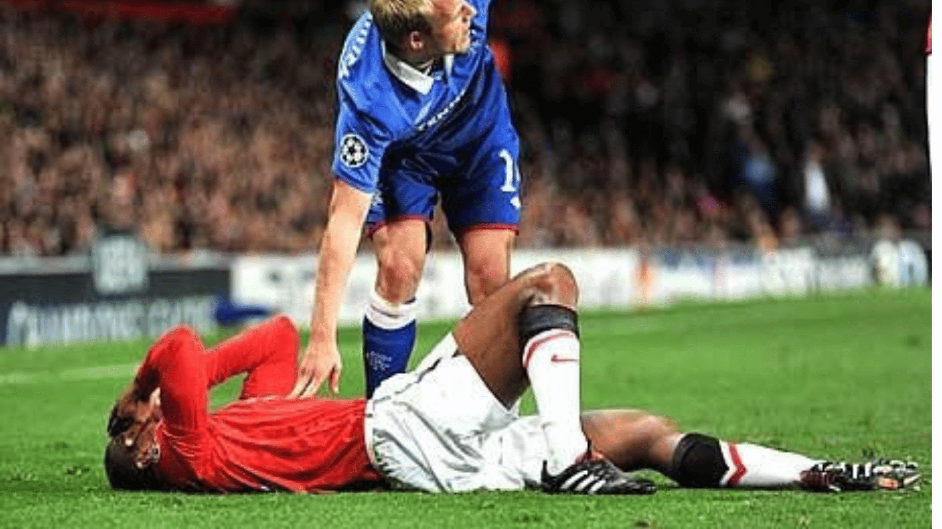 soccer player in blue stands over an injured player in red lying on the grass holding his head during a match