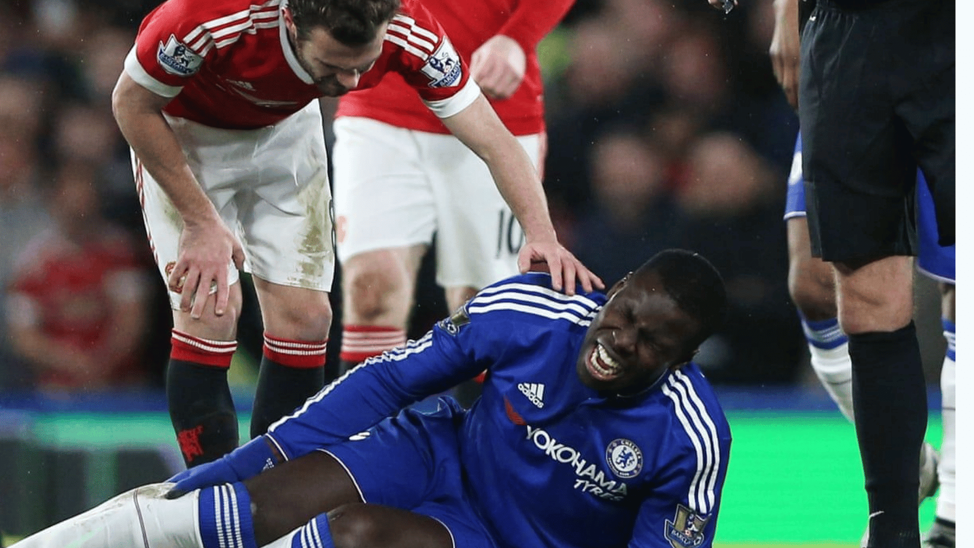 soccer player in blue uniform sits on the field grimacing as opponent in red uniform checks on him during a match