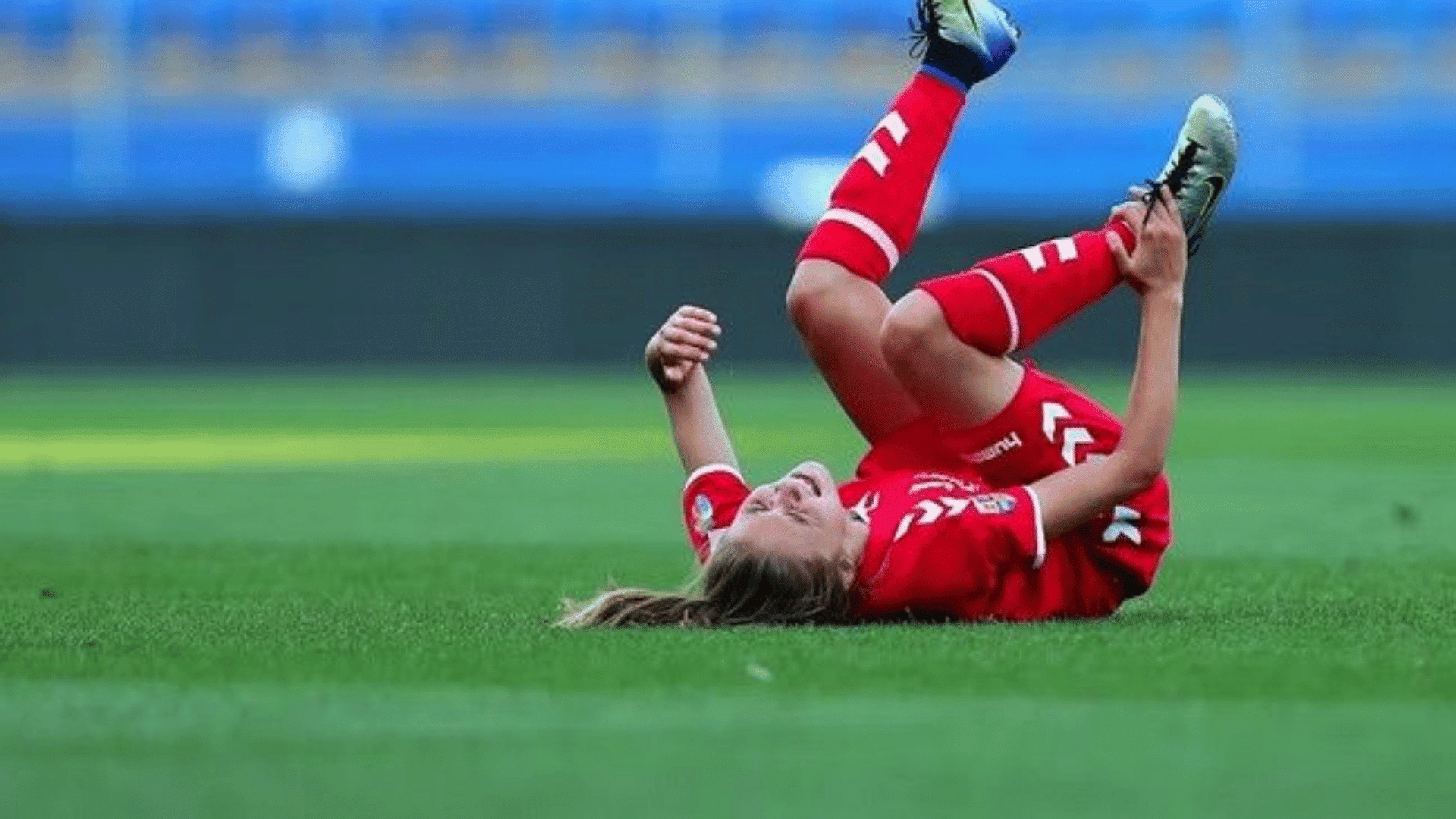 soccer player in red kit lying on grass on back with legs raised holding foot in possible pain during match
