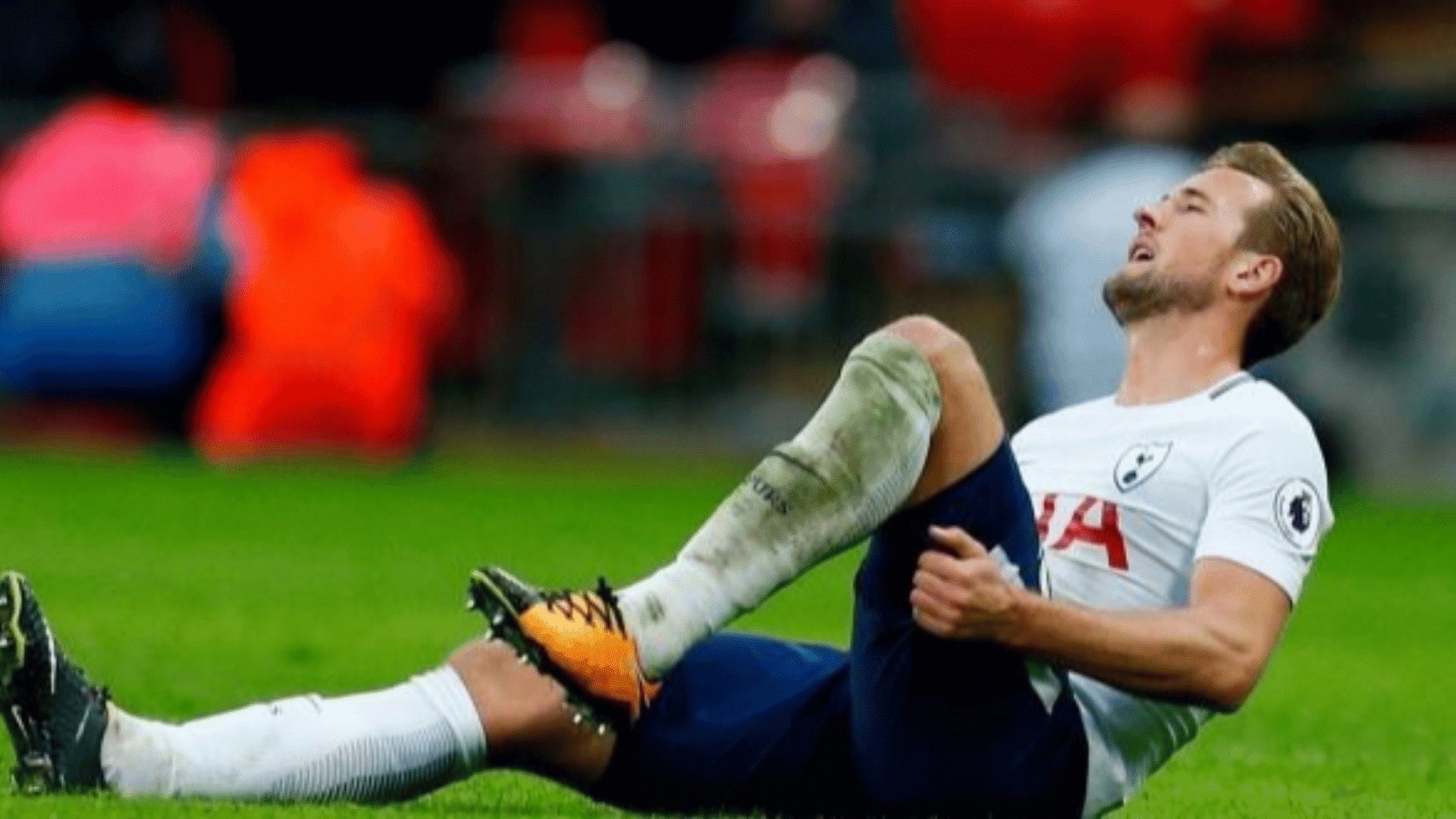 soccer player in white kit sitting on grass holding injured leg in pain grimacing during stadium match blurred crowd