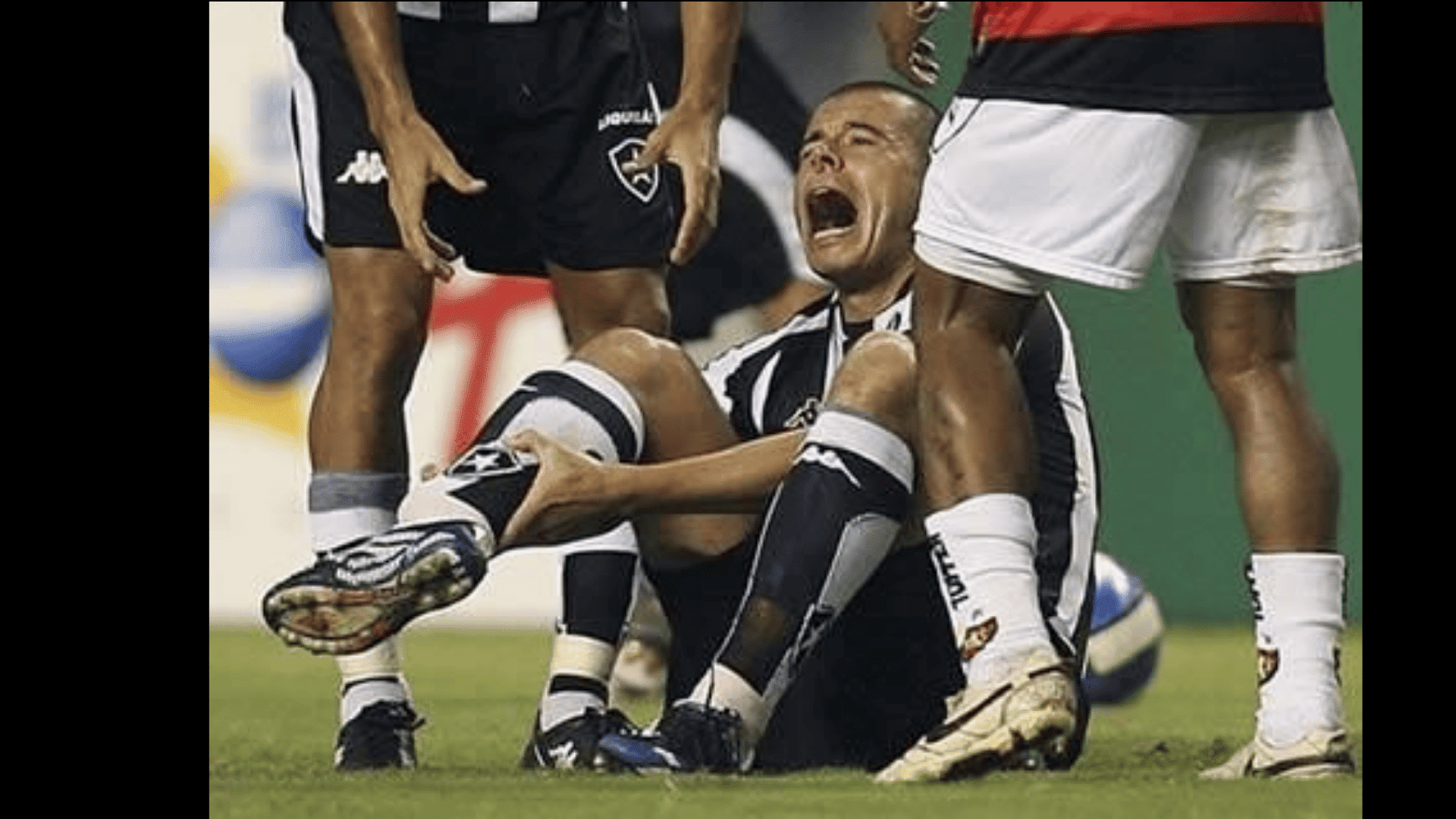 soccer player sits on the grass crying and holding his leg while teammates stand nearby during a match
