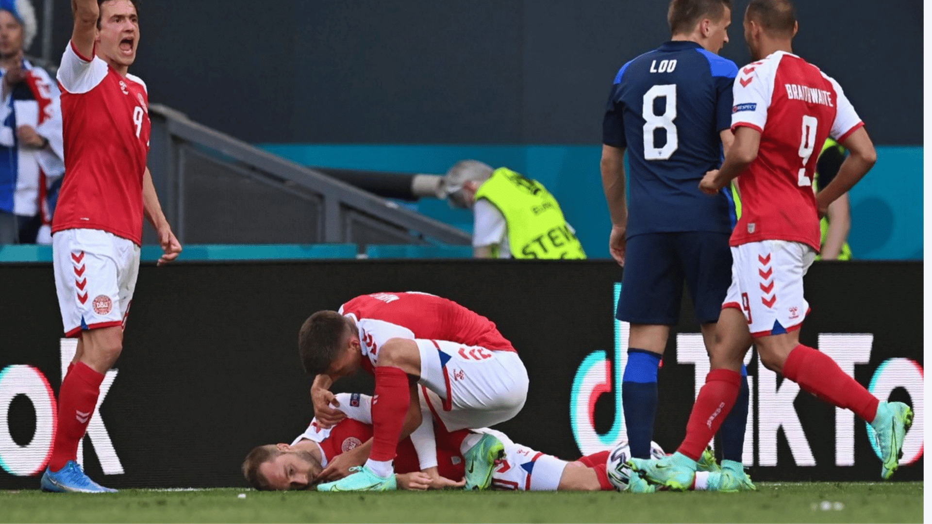 soccer players gather as one player lies injured on the grass while a teammate bends over to check on him