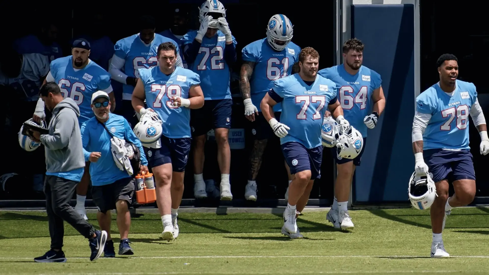 tennessee titans players in blue practice jerseys jog from a dark tunnel onto a sunny grass football field