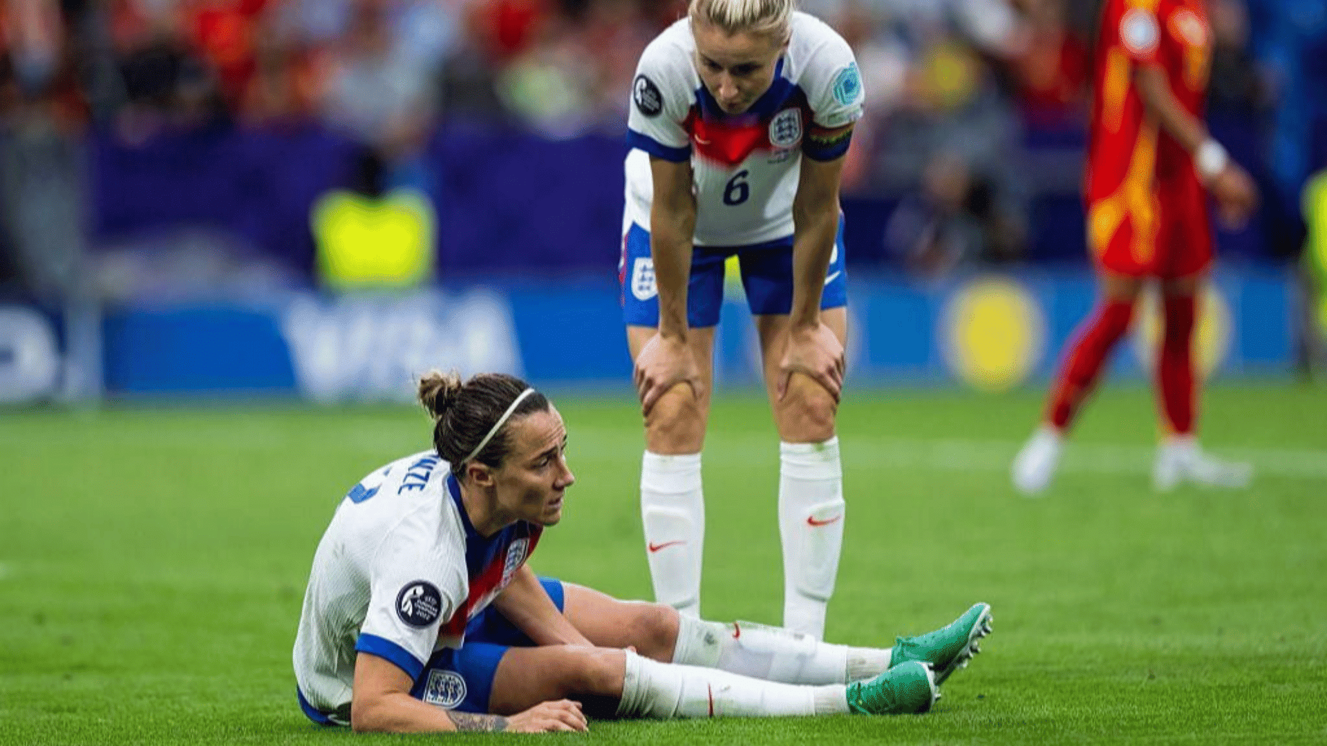 two england women's soccer players one sitting on grass holding leg in pain while teammate stands looking concerned