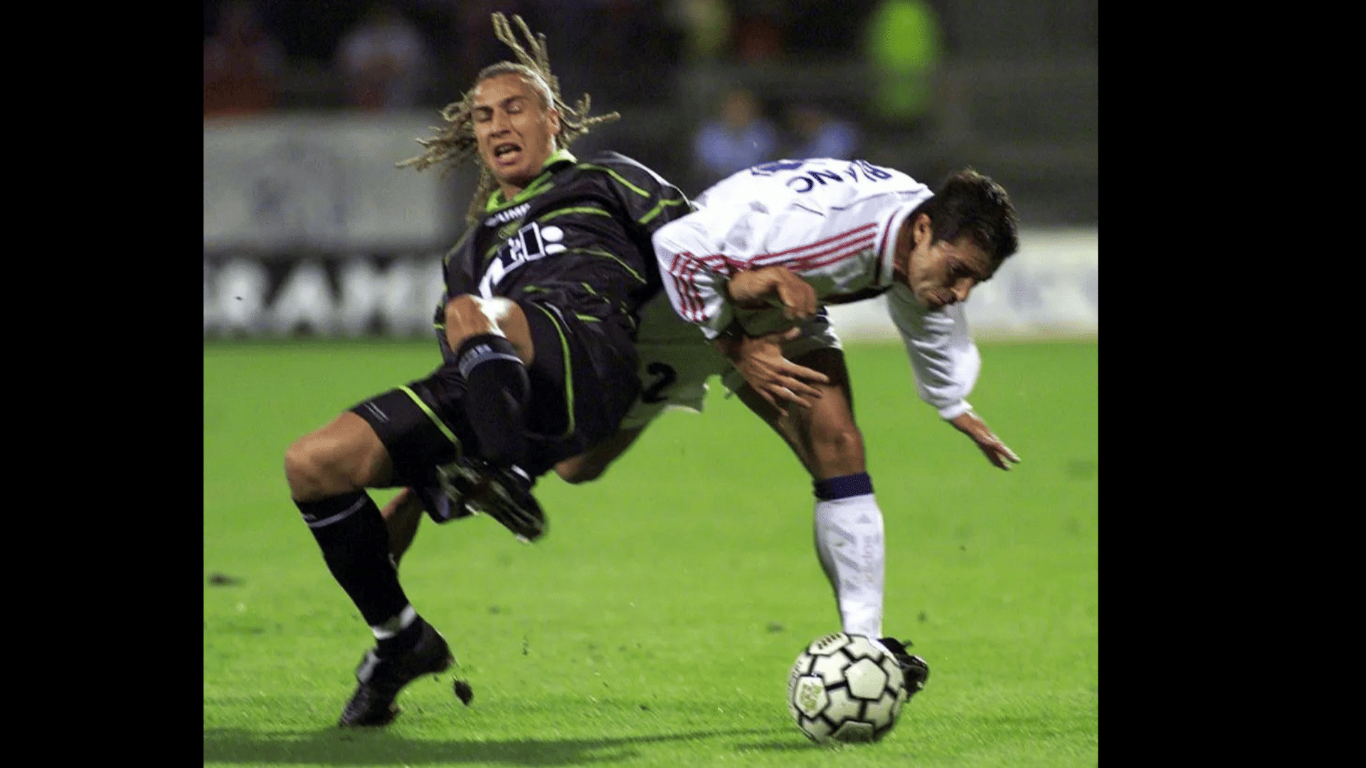 two football players clash near the ball as one stumbles forward and the other falls back during a night match