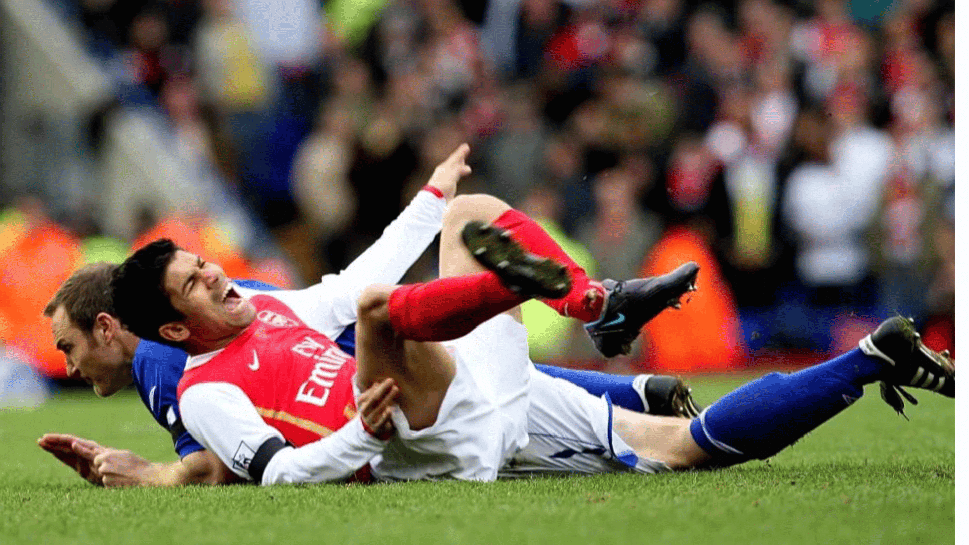 two football players fall to the grass during a hard tackle with a blurred crowd watching from the stands