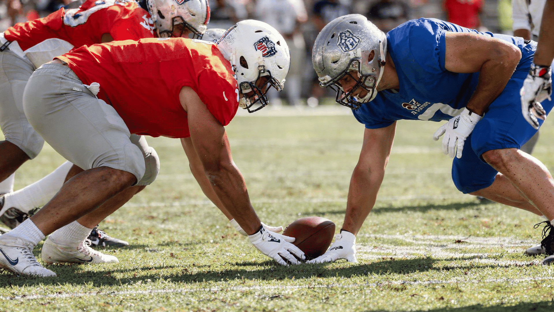 two football players in red and blue uniforms are facing each other in a ready stance over a football on a grassy field